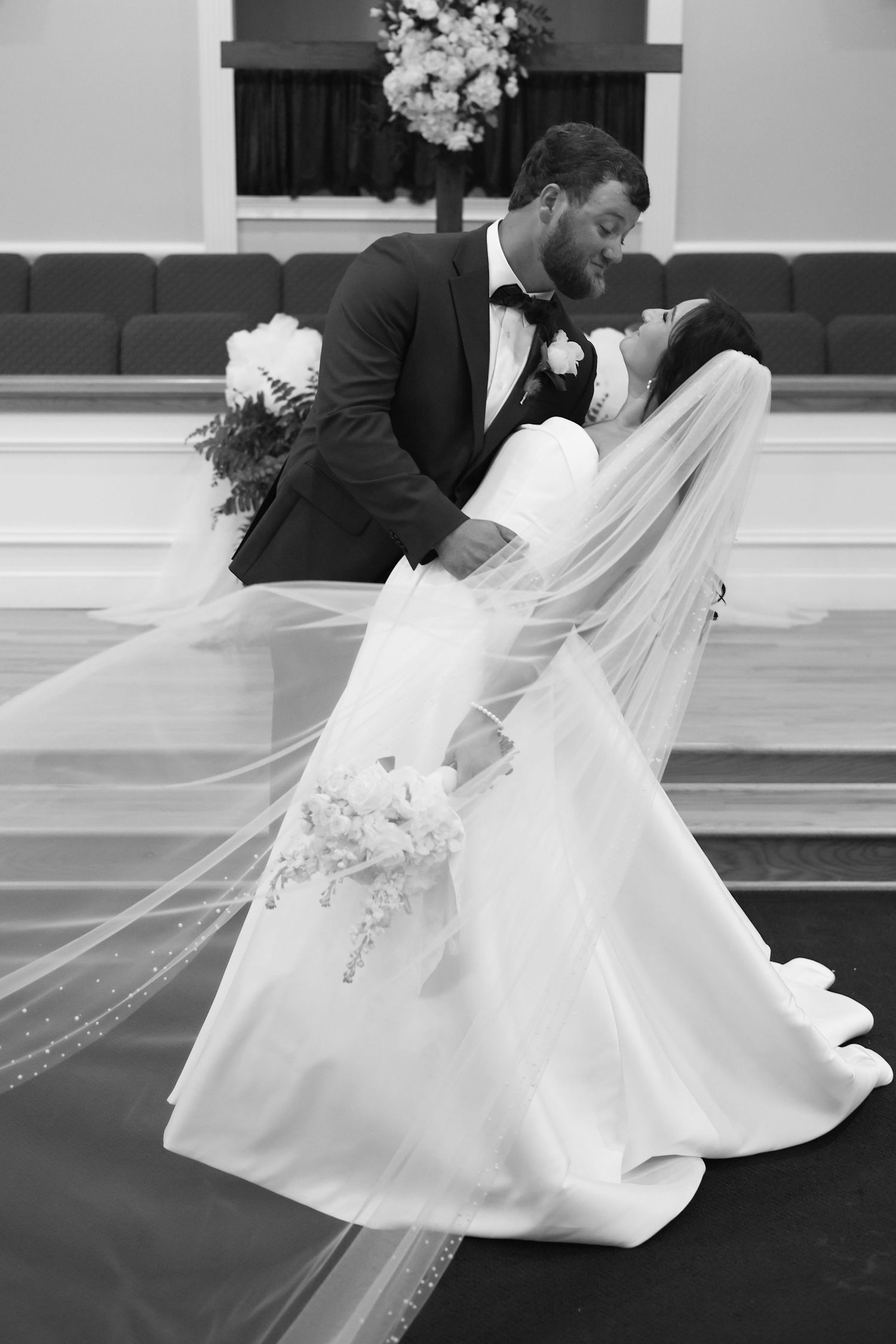 Black and white wedding photo of groom dipping bride for a kiss in church, her veil flowing dramatically.