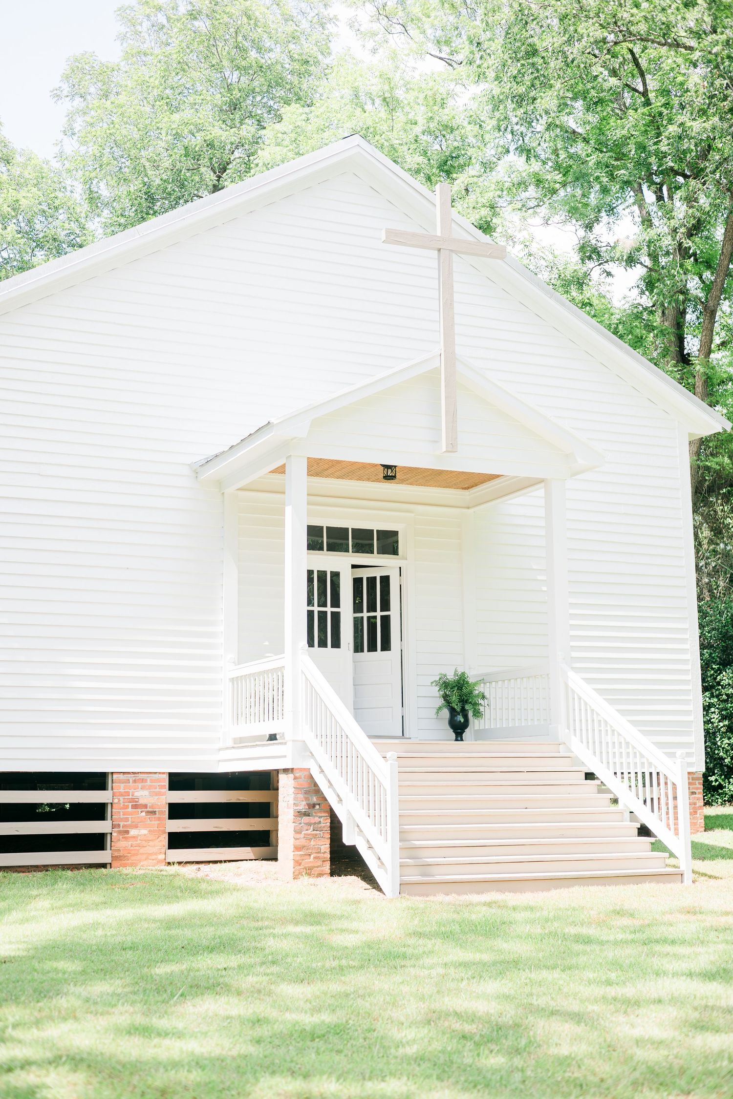 Charming white wooden chapel with cross, front porch, and steps surrounded by green trees on a sunny day.