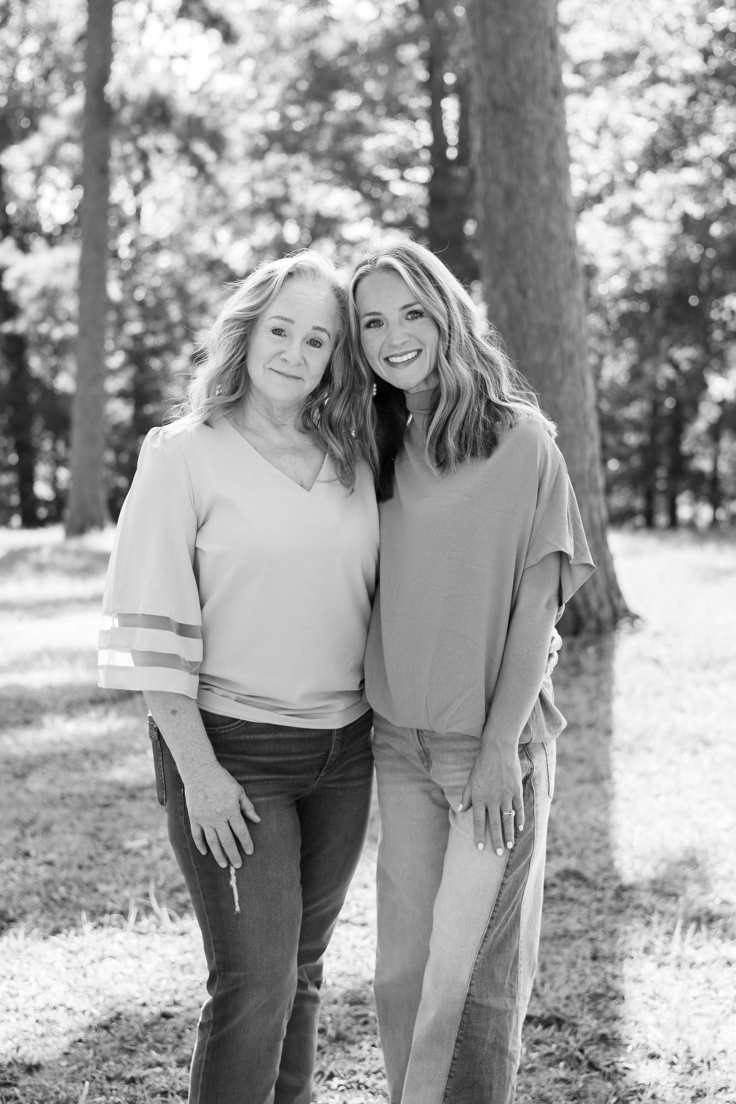 Two smiling women pose together outdoors among tall trees in a black and white portrait photo.