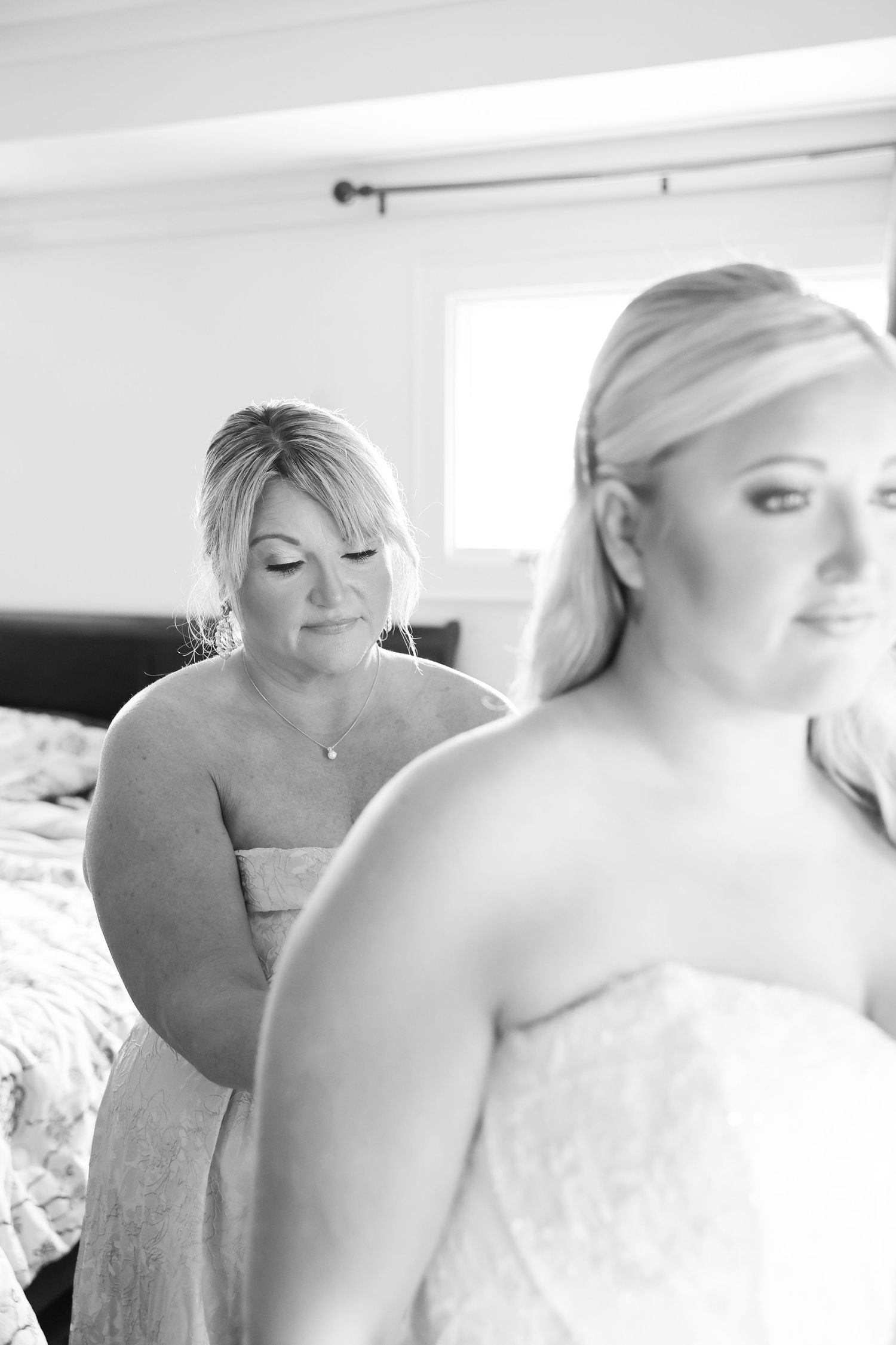 Black and white photo of a bride being helped into her wedding dress by a woman in a strapless gown in a bright bedroom.