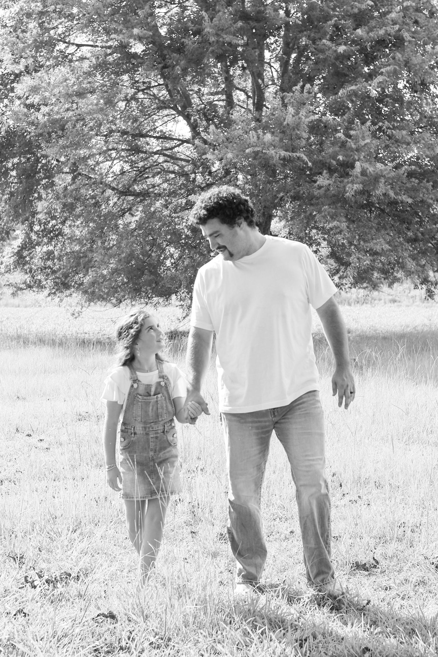 Black and white photo of a father and young daughter holding hands while walking together in a grassy field.