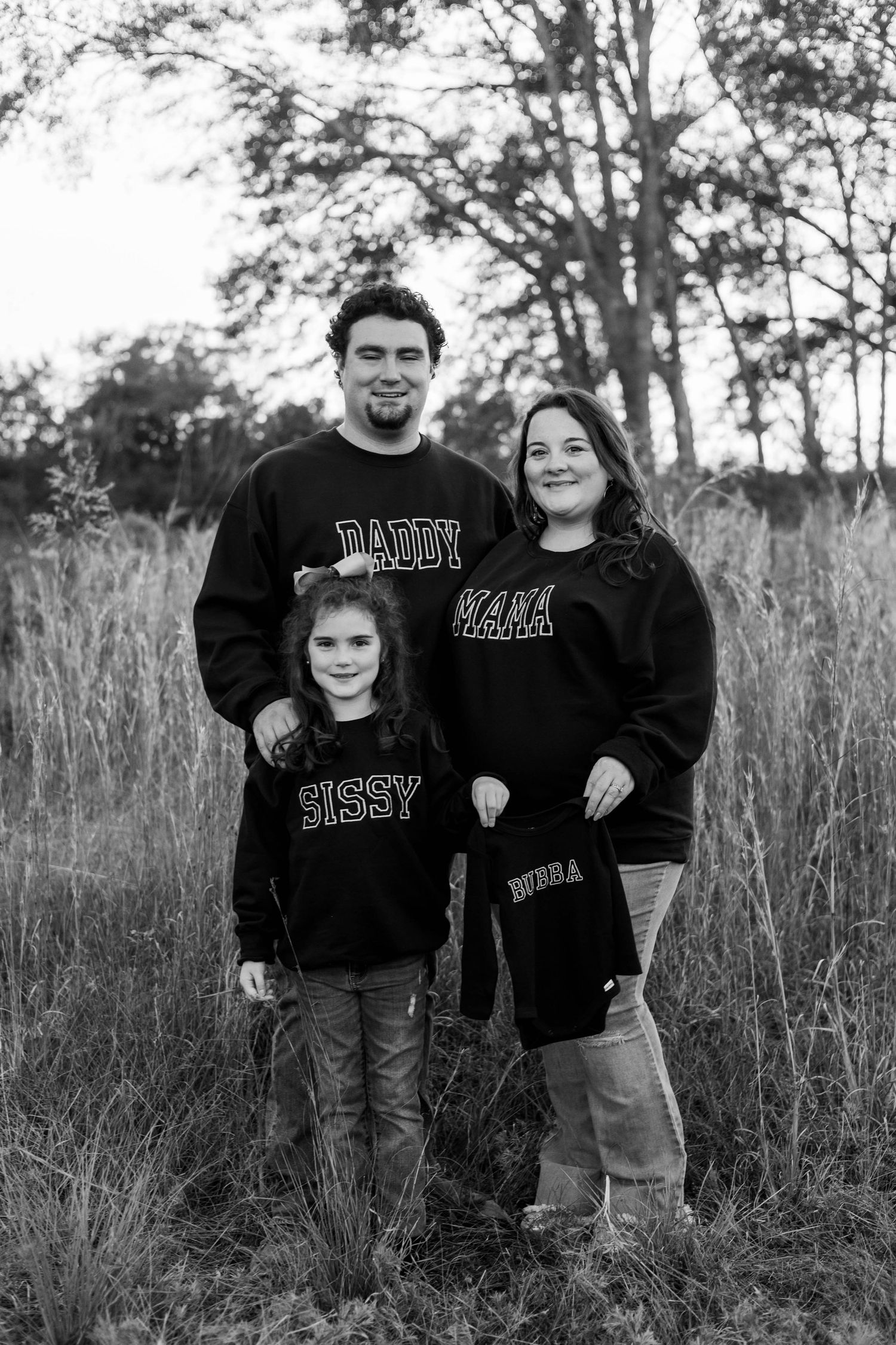 Black and white family photo with matching sweatshirts reading Daddy, Mama, Sissy in a field.