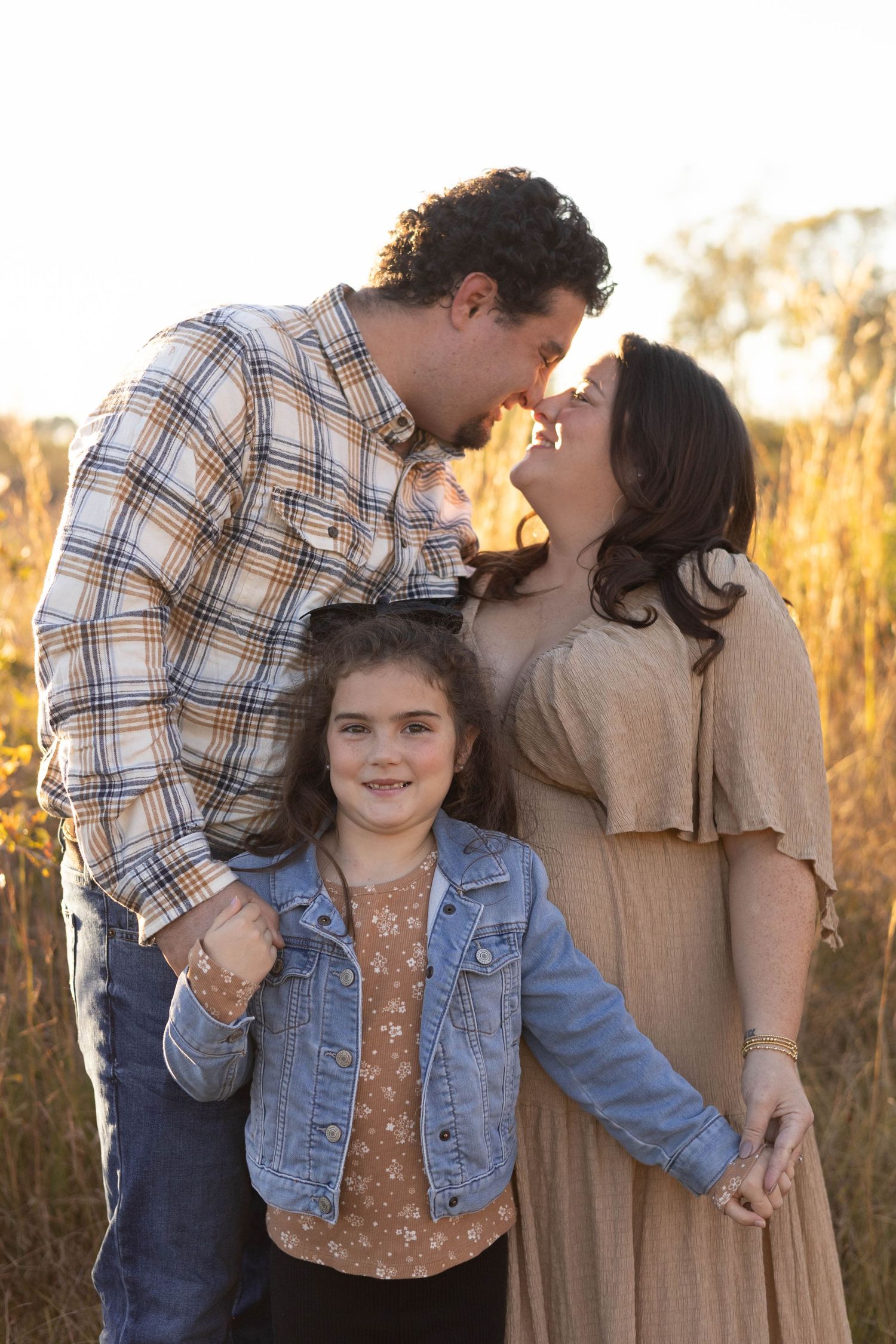 A family of three poses outdoors at golden hour, parents sharing a kiss while their young daughter smiles in a denim jacket.