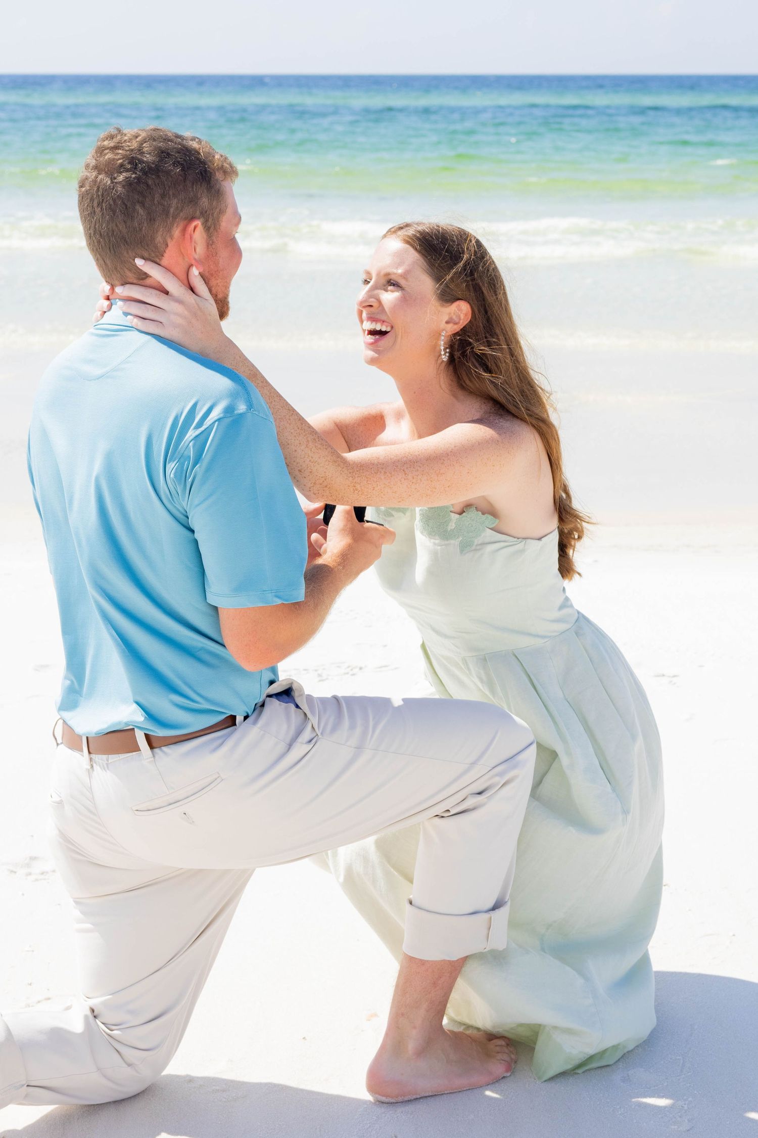 Man on one knee proposing to emotional woman in mint dress on a sunny beach by the ocean.