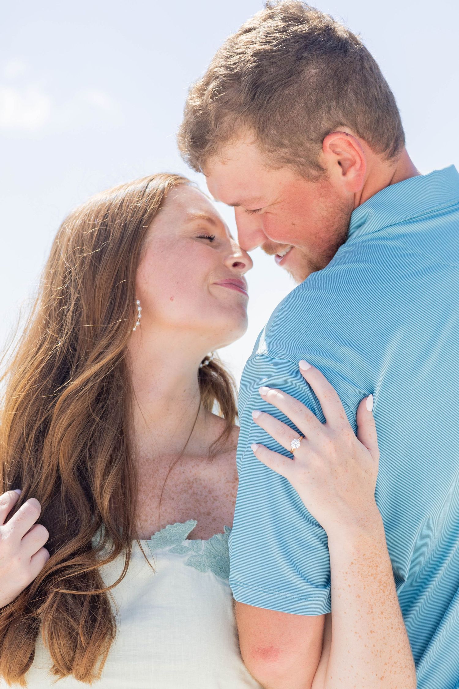Smiling couple nearly kissing with womans engagement ring visible as she embraces man in blue shirt.