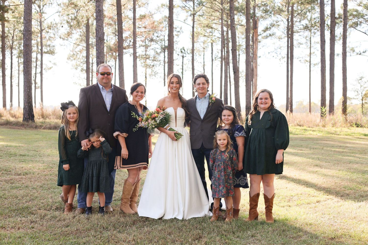 Large wedding party poses together among tall pine trees at sunset.