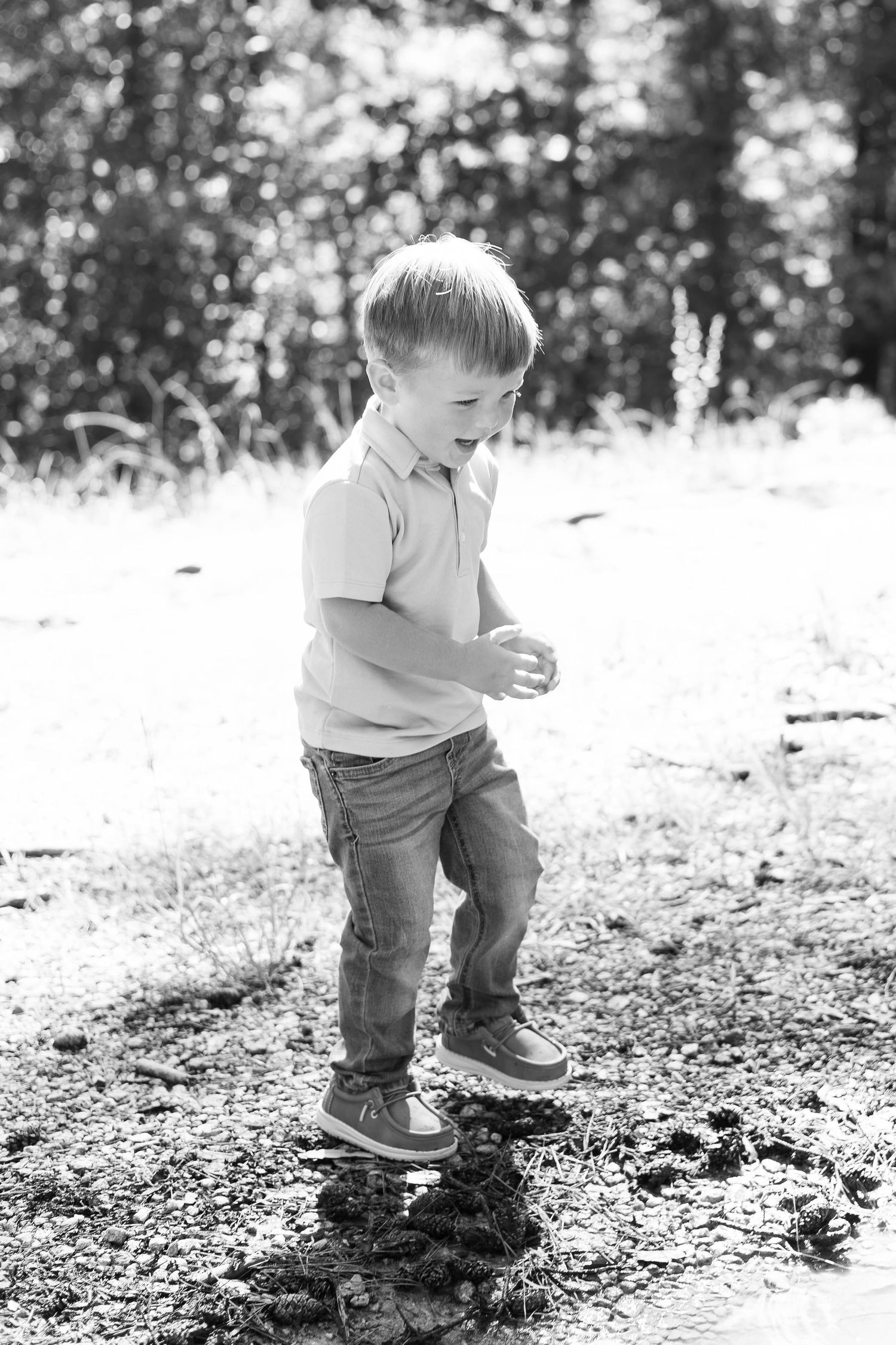 Black and white photo of a young boy playing in dirt outdoors in a sunny forest clearing, wearing jeans and a polo shirt.