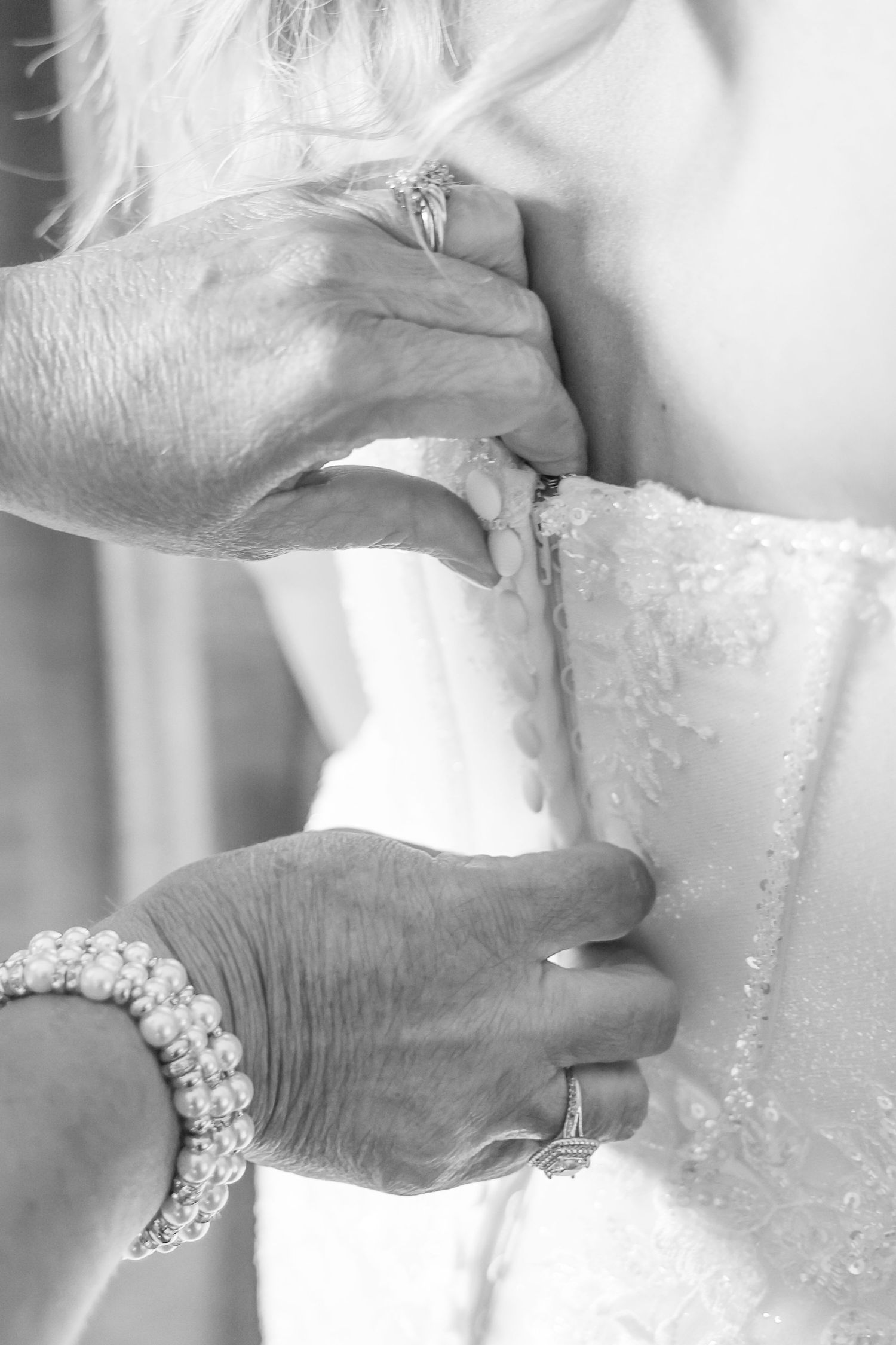 Black and white photo of hands fastening a bridal gown, with pearl bracelet and rings visible.