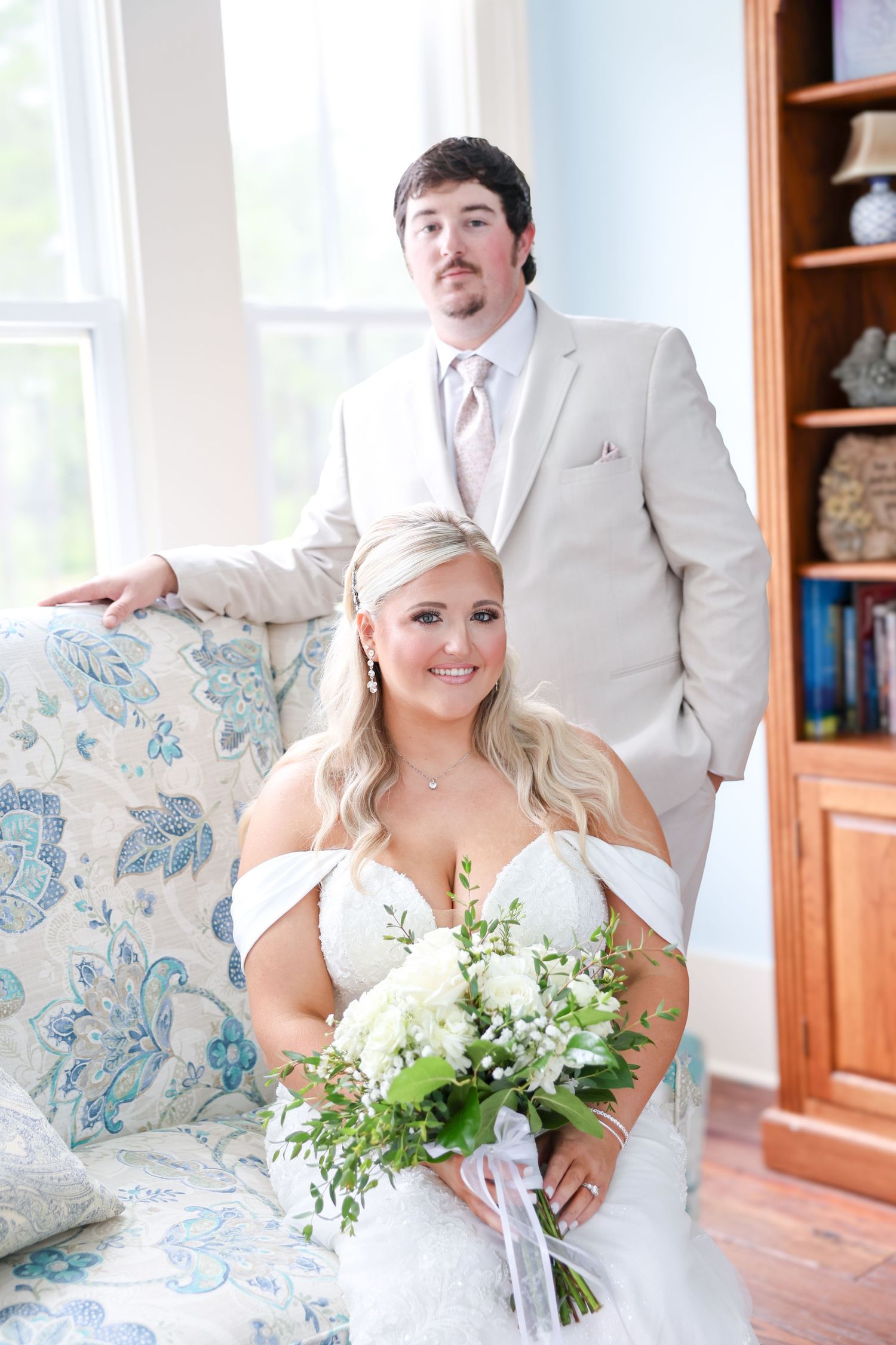 Bride and groom pose elegantly in a sunlit room, bride holding white floral bouquet in lace gown, groom in tan suit.