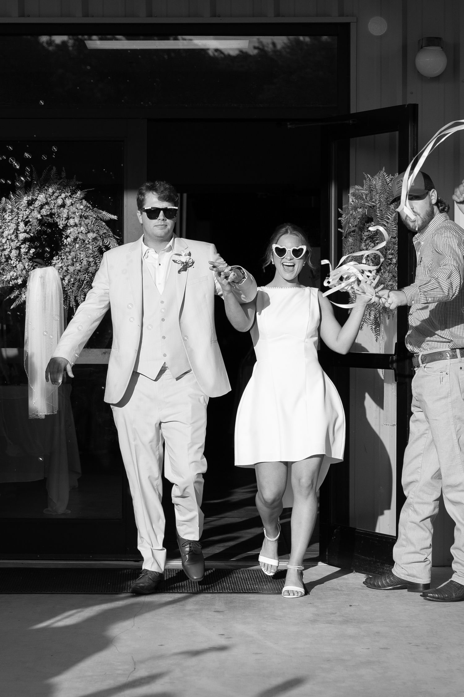 Newlywed couple in sunglasses laughing as they exit wedding venue doors in black and white photo.