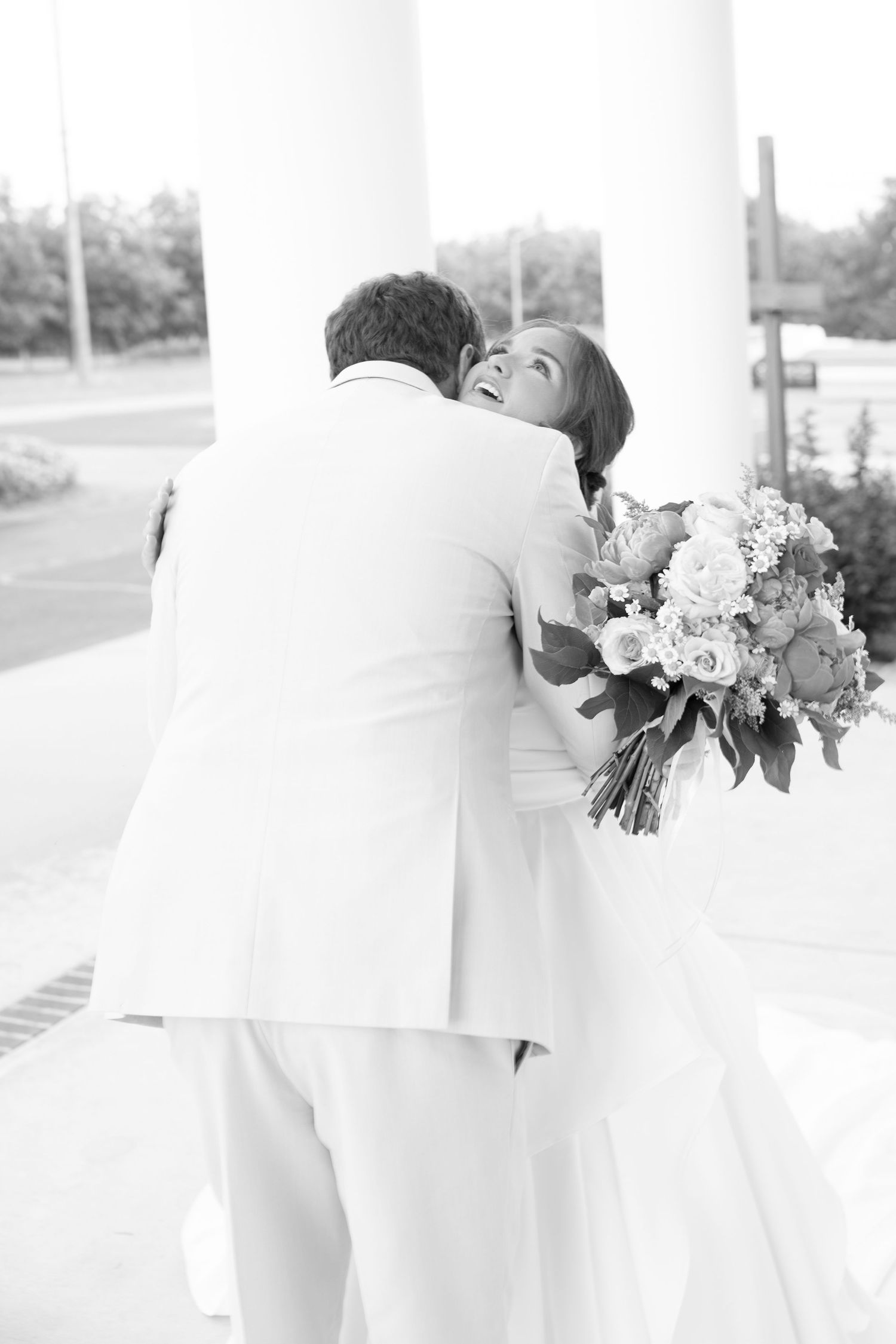 Black and white photo of a bride holding a floral bouquet embracing her groom in a white suit near elegant columns.