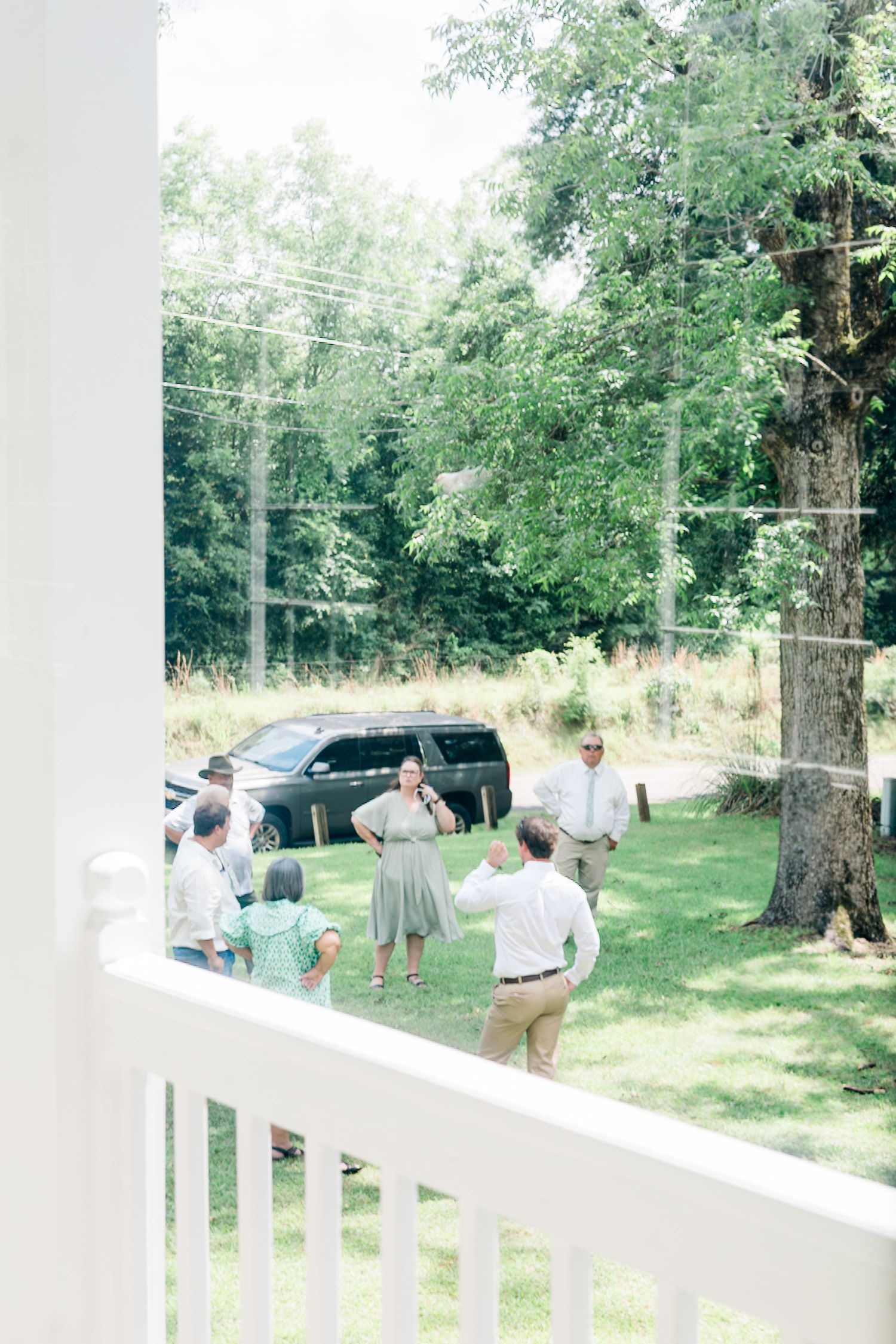 Wedding guests mingling outdoors on green lawn viewed through white porch railing.