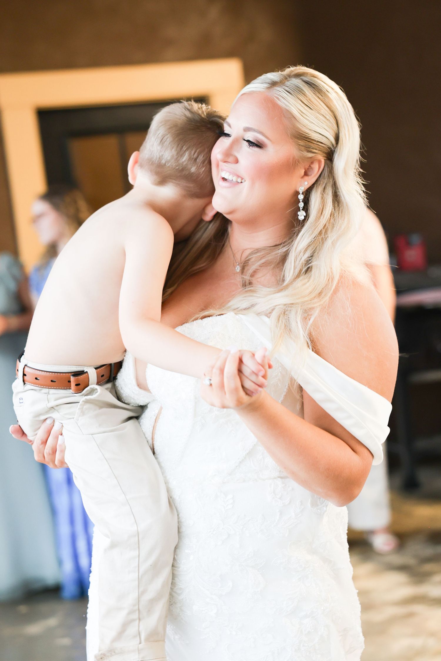 A radiant bride in a white off-shoulder gown holds a young boy who whispers in her ear at the reception.