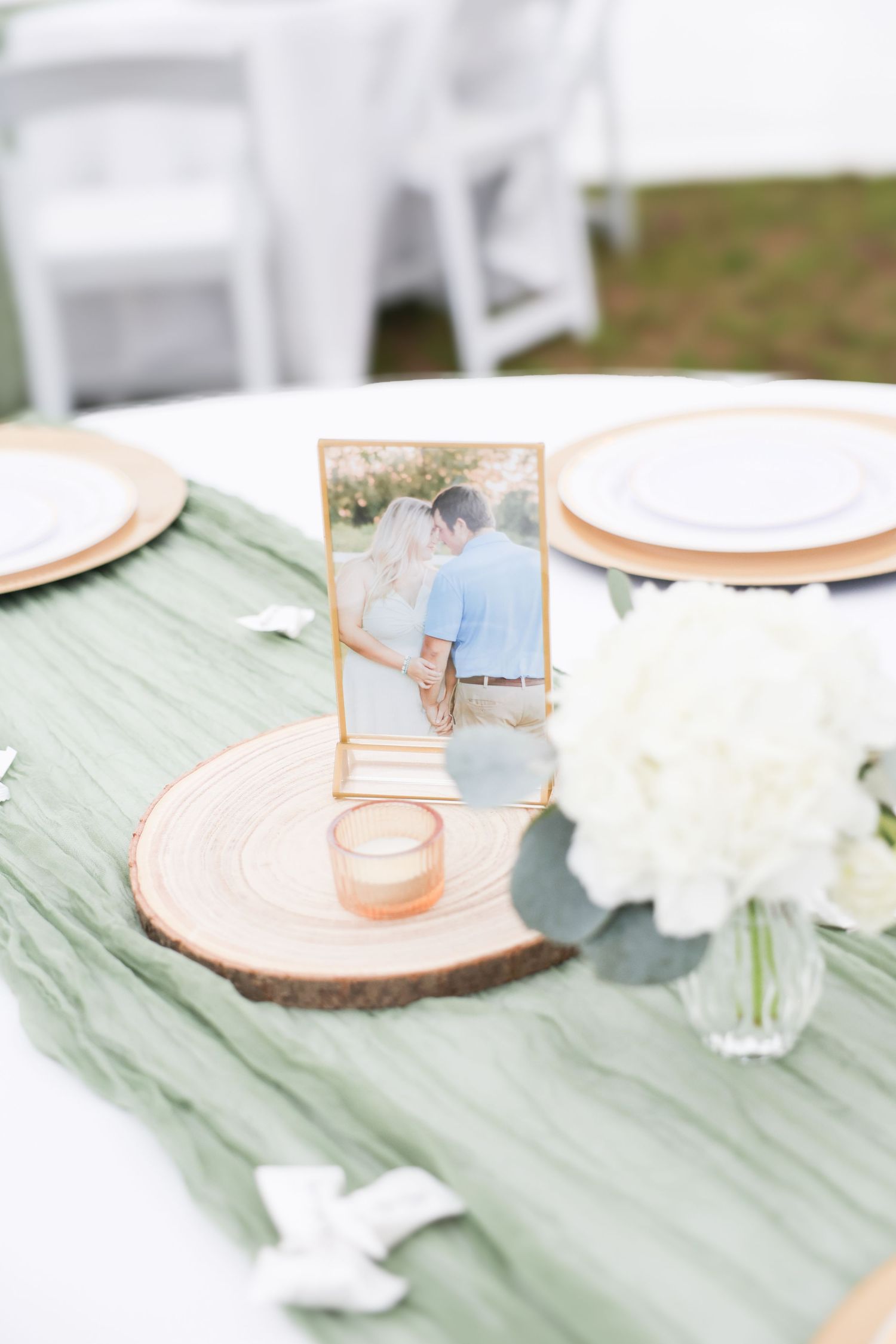 Elegant wedding reception table with sage green runner, wood slice centerpiece, couple photo, and white hydrangeas.