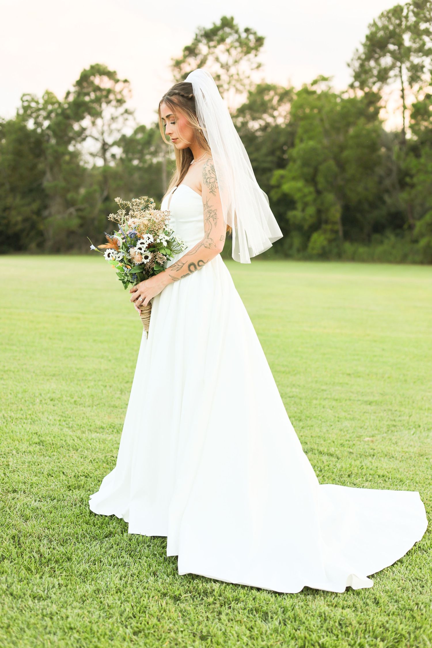 A bride in a flowing white gown poses gracefully on a sunlit grassy field.