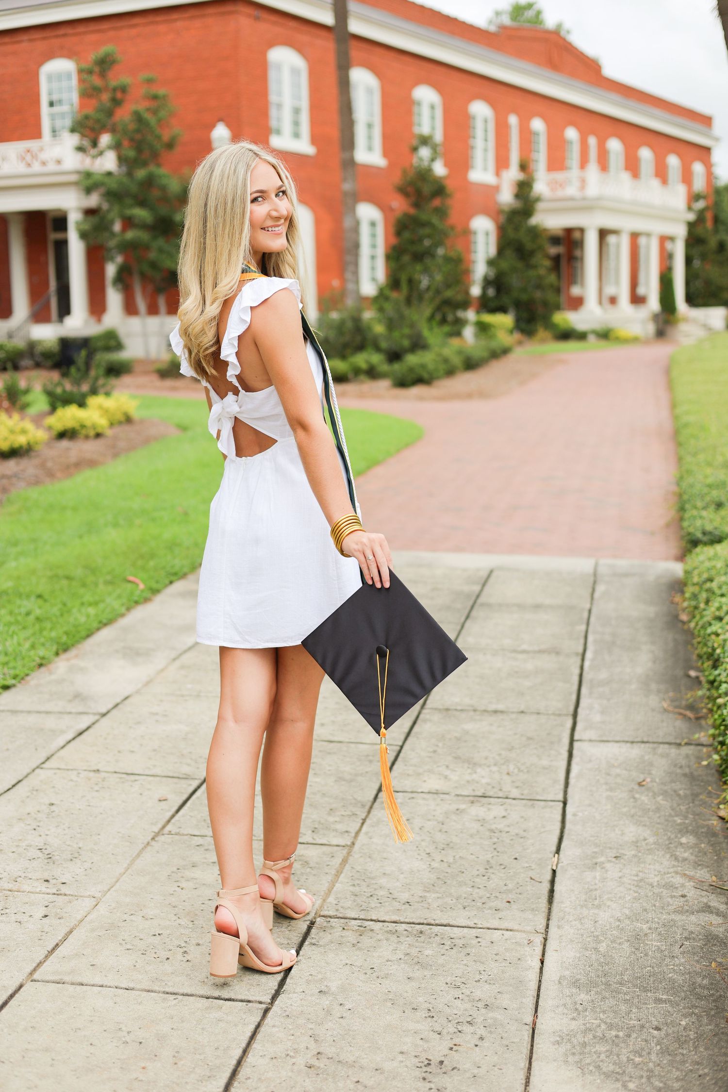 Graduate in white dress poses with cap near red brick building on stone pathway.