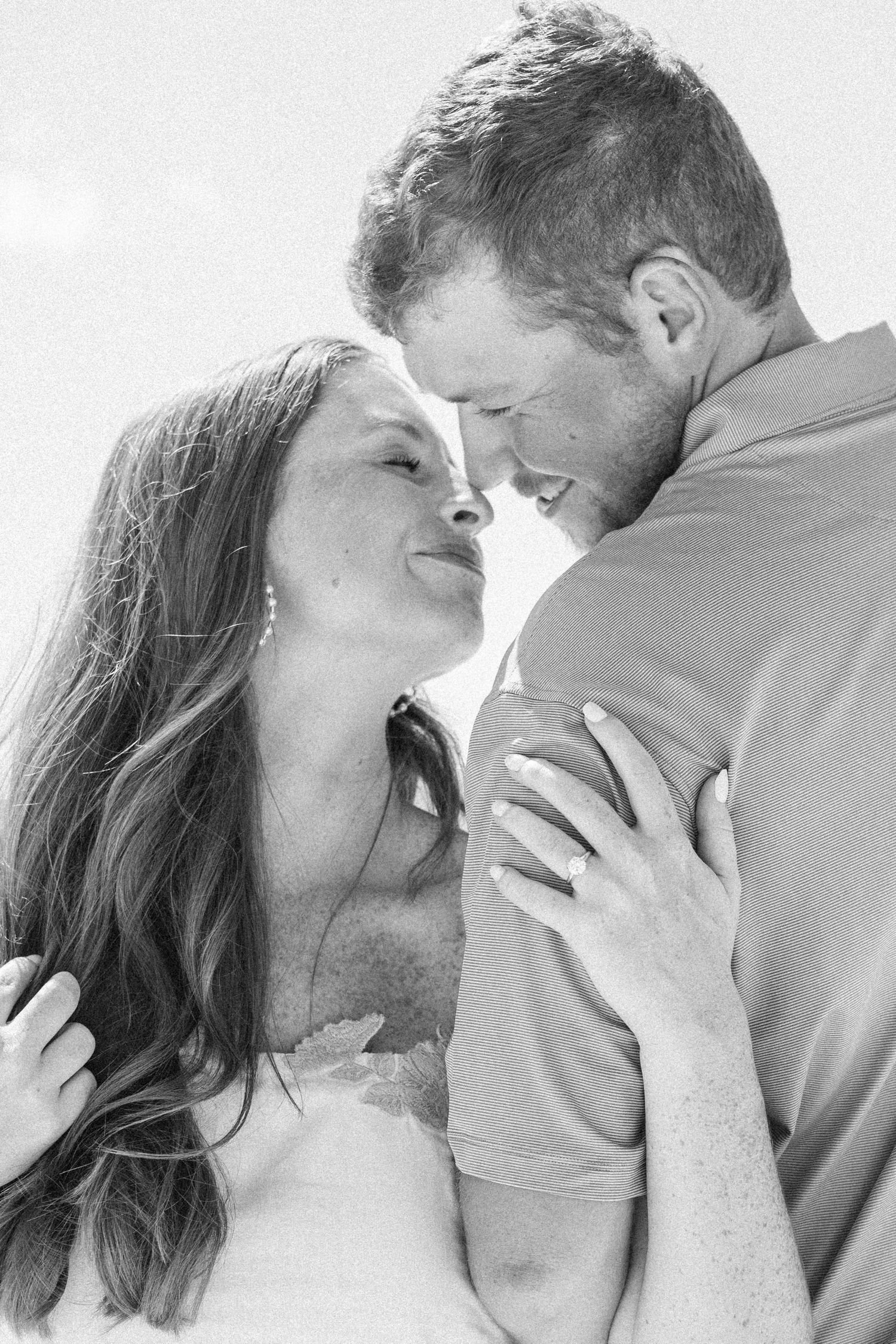 Black and white romantic beach portrait of smiling couple about to kiss, woman wearing engagement ring embracing man.