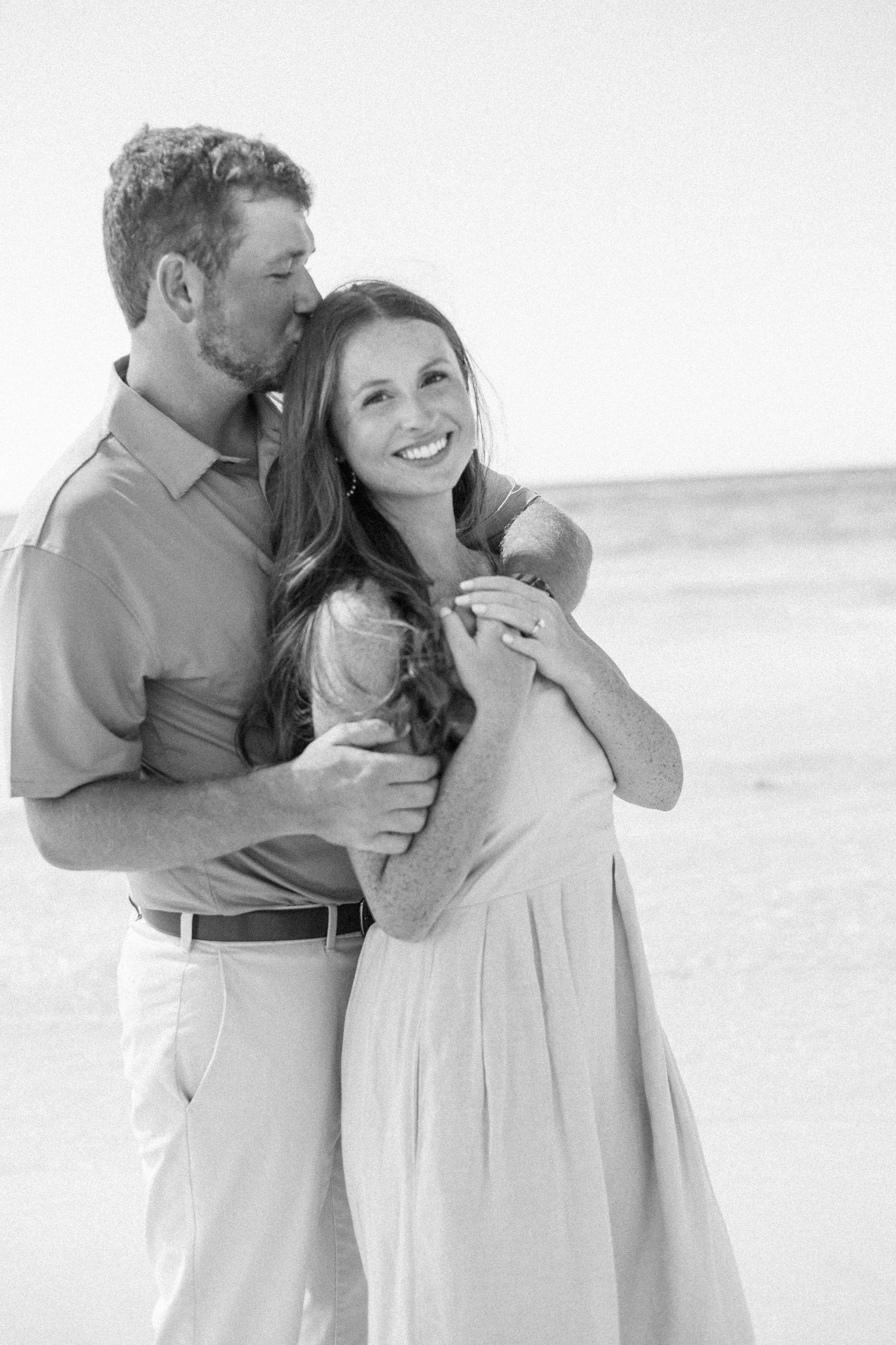 Man tenderly kissing woman on cheek while embracing her from behind during beach engagement session.