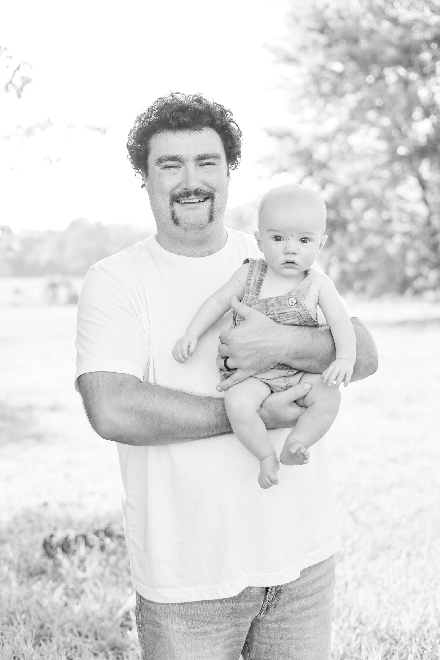 Black and white photo of a smiling man with a mustache holding a baby in overalls outdoors.