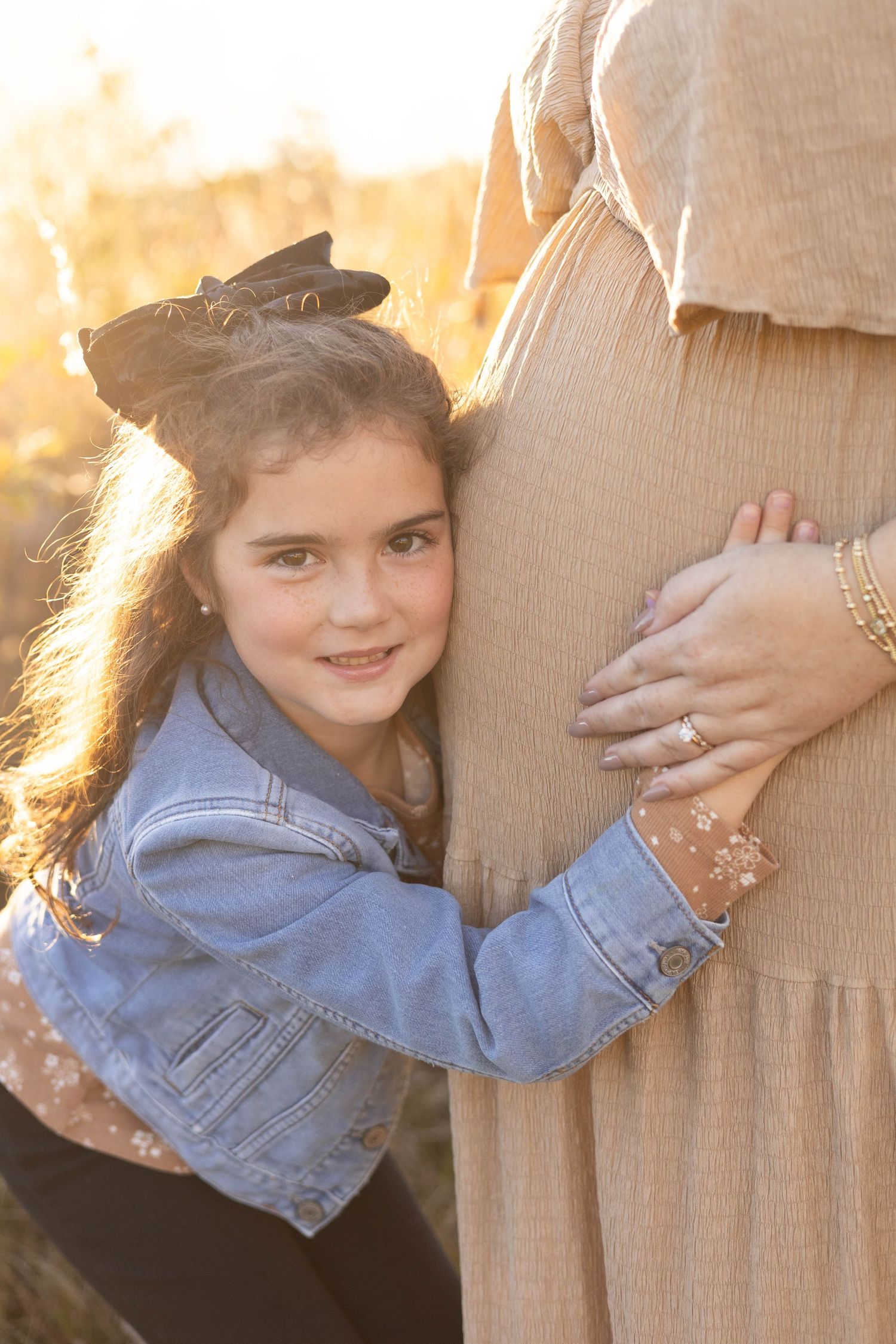 Young girl in denim jacket hugging pregnant woman's belly in golden sunset light.