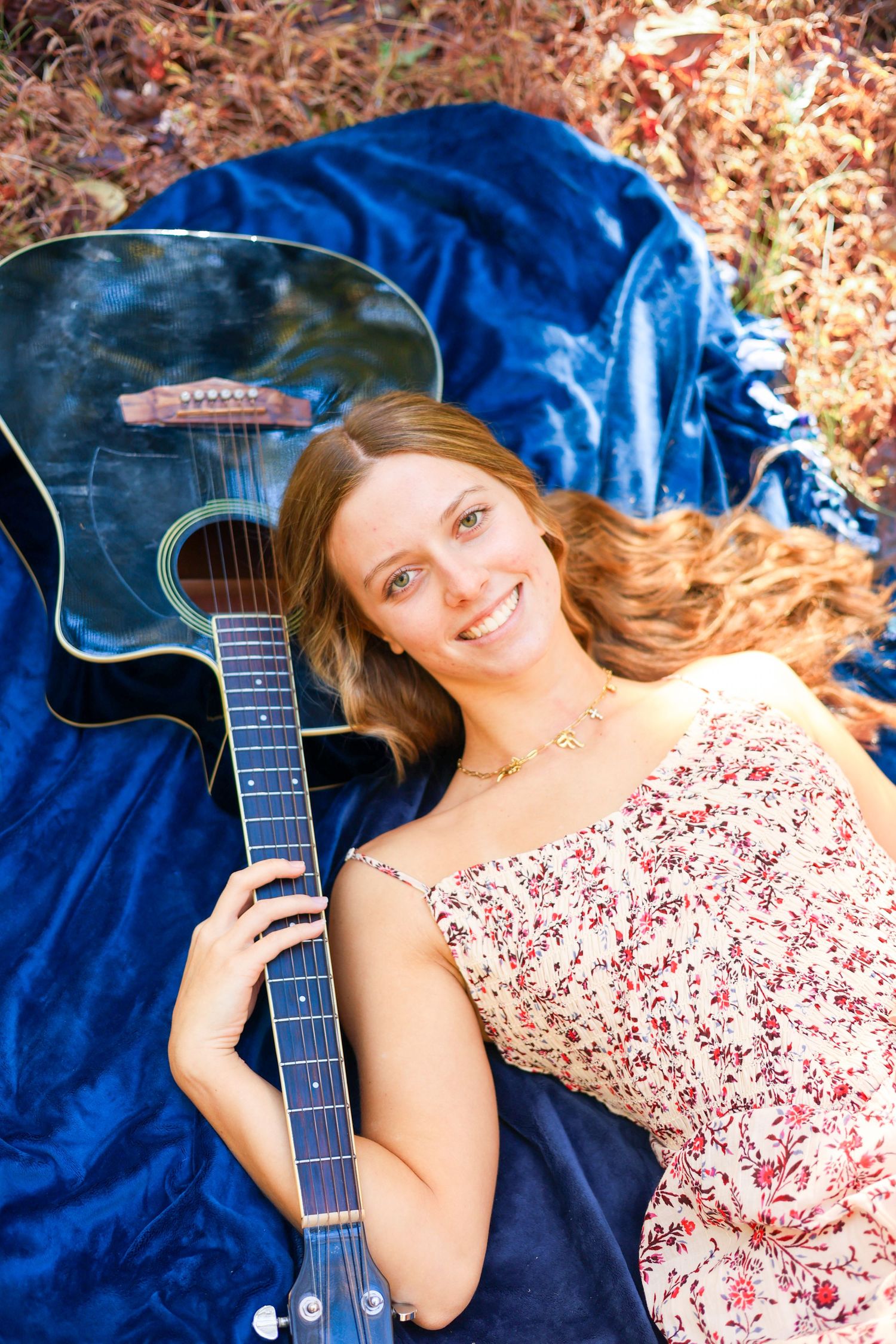 Person in floral dress poses with acoustic guitar on blue blanket outdoors.