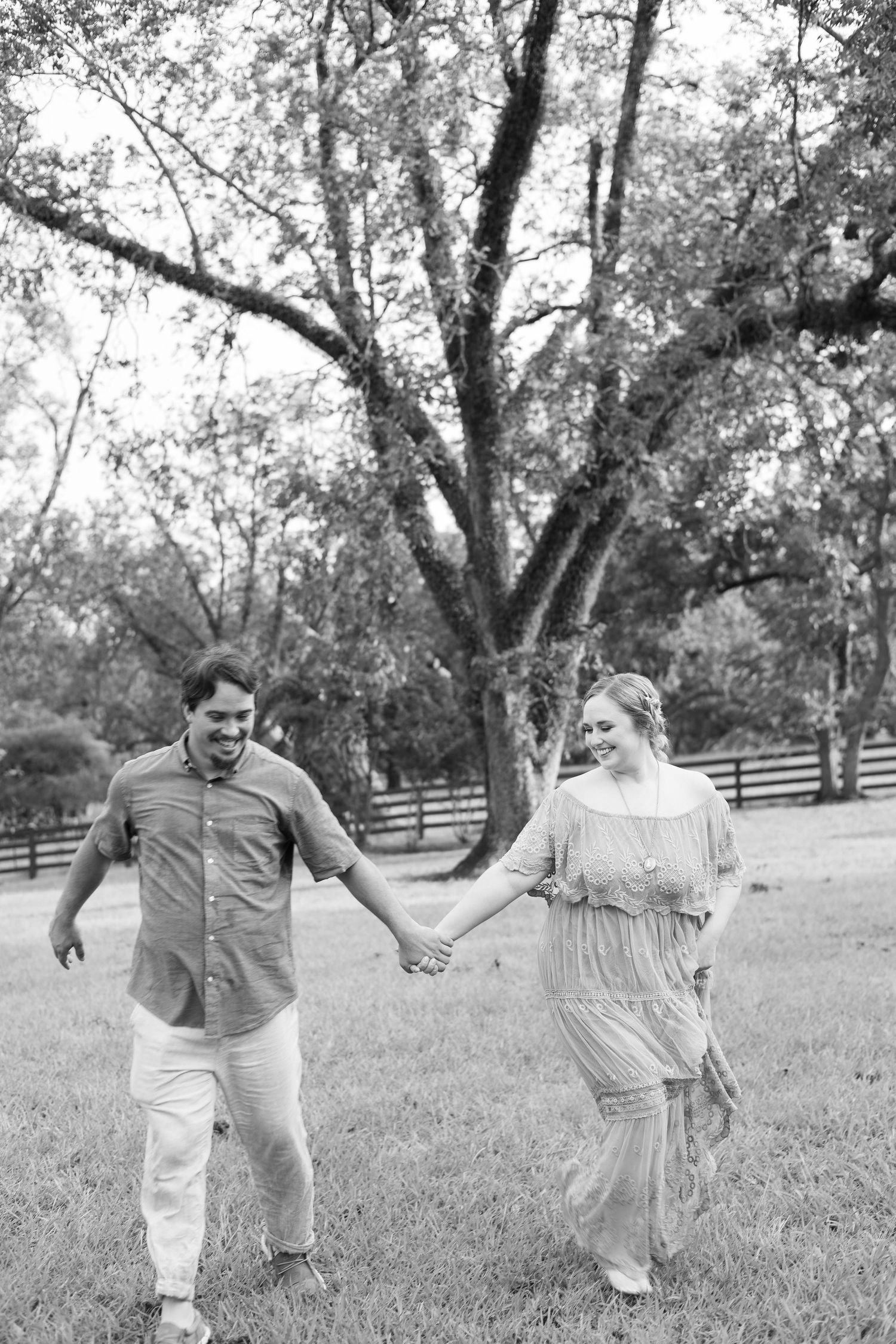 Black and white photo of a happy couple holding hands and walking through a grassy park with trees and a fence.