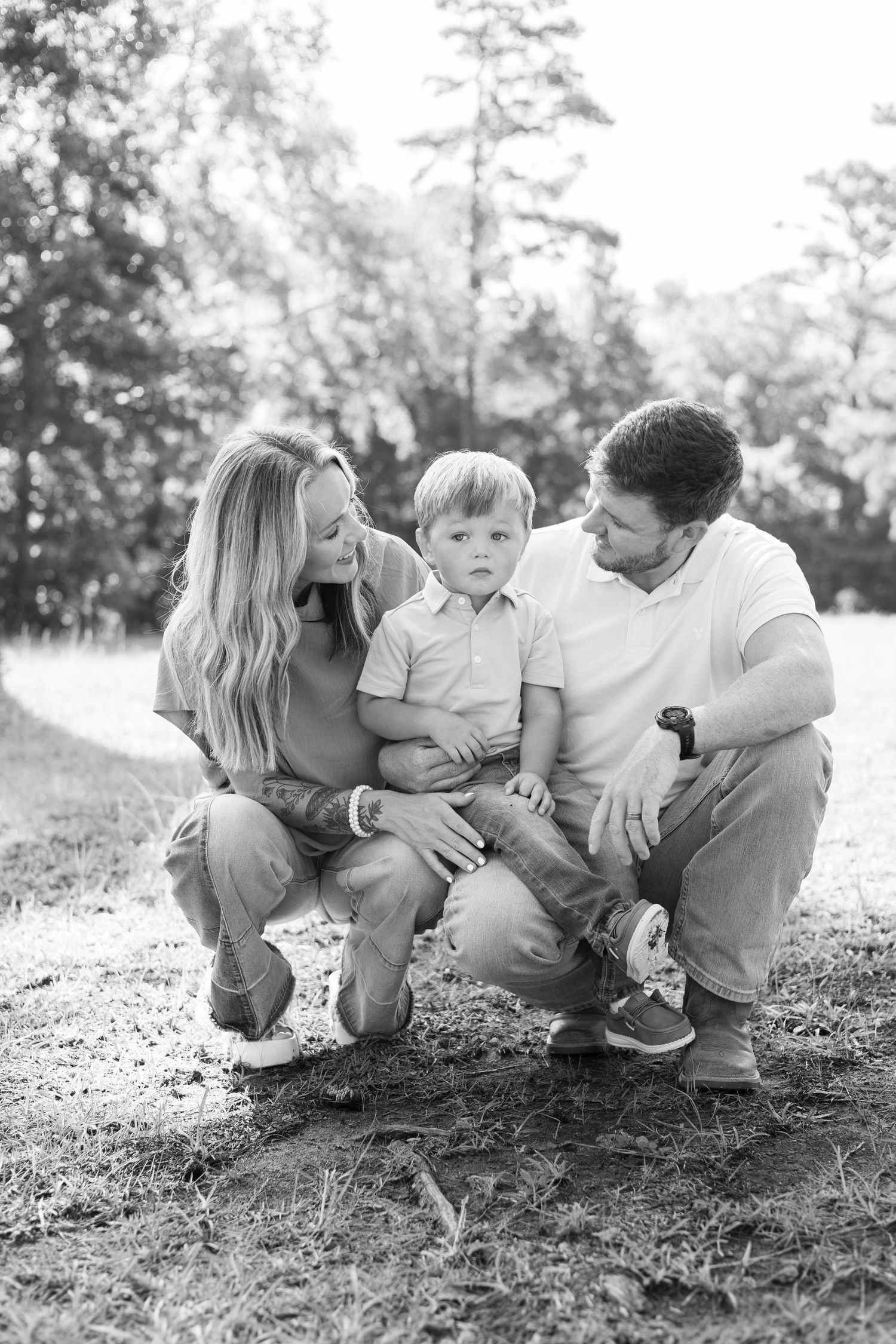 Black and white family portrait of parents crouching with young toddler son outdoors in a park setting.