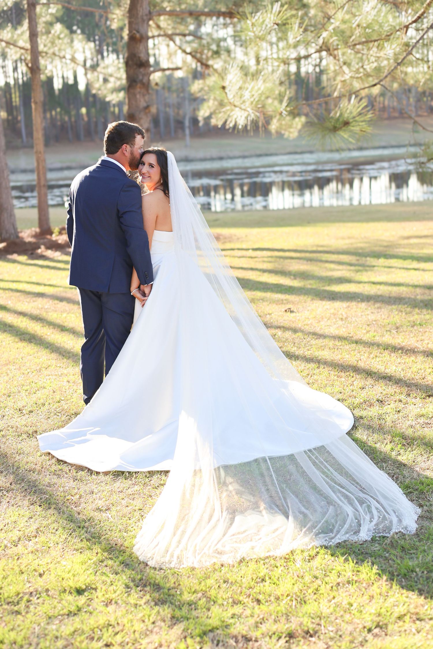 Romantic wedding couple embraces in a sunlit park with flowing white dress and navy suit.
