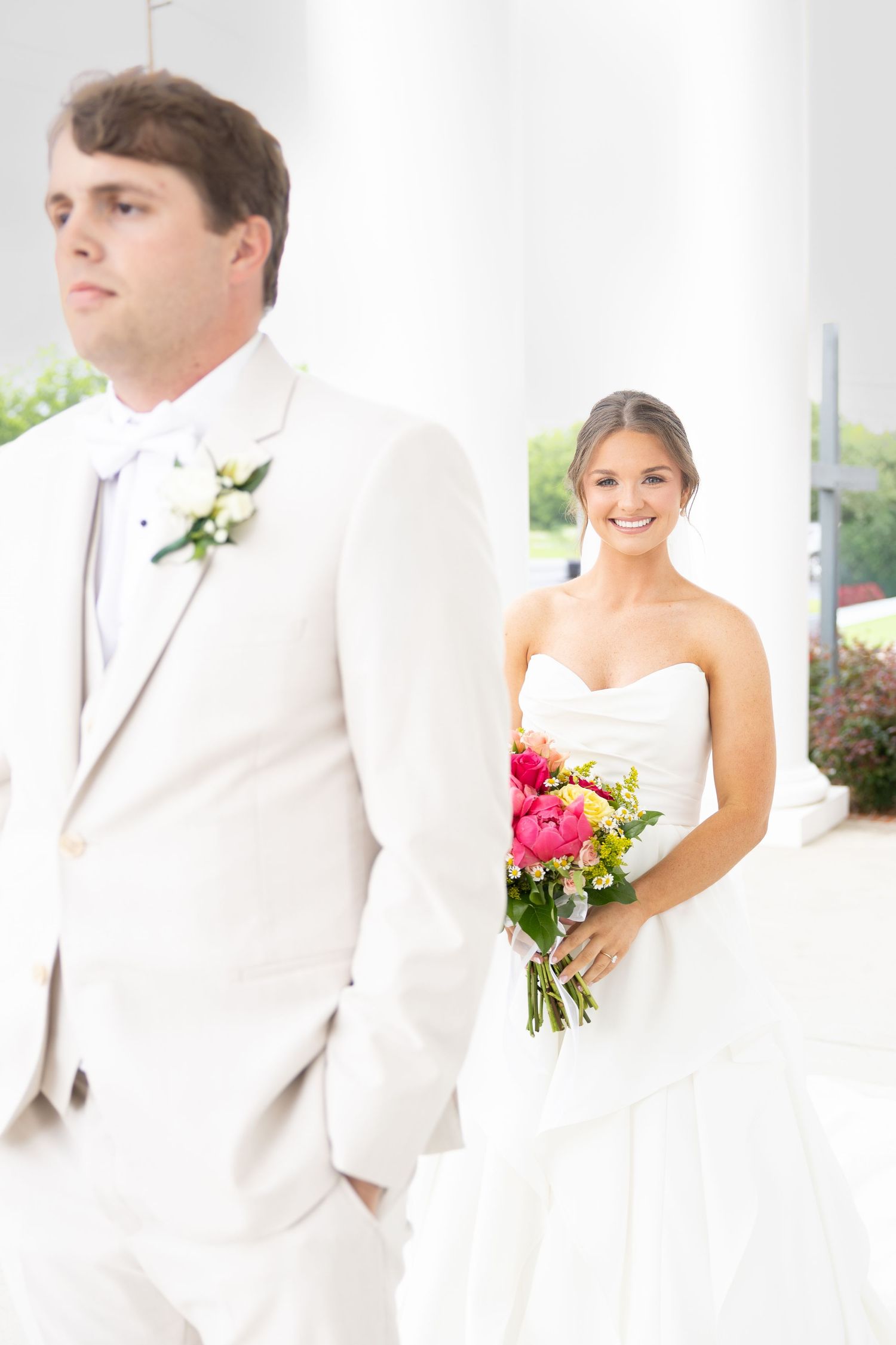 Smiling bride in strapless gown holds colorful bouquet behind groom in white tux at outdoor venue.
