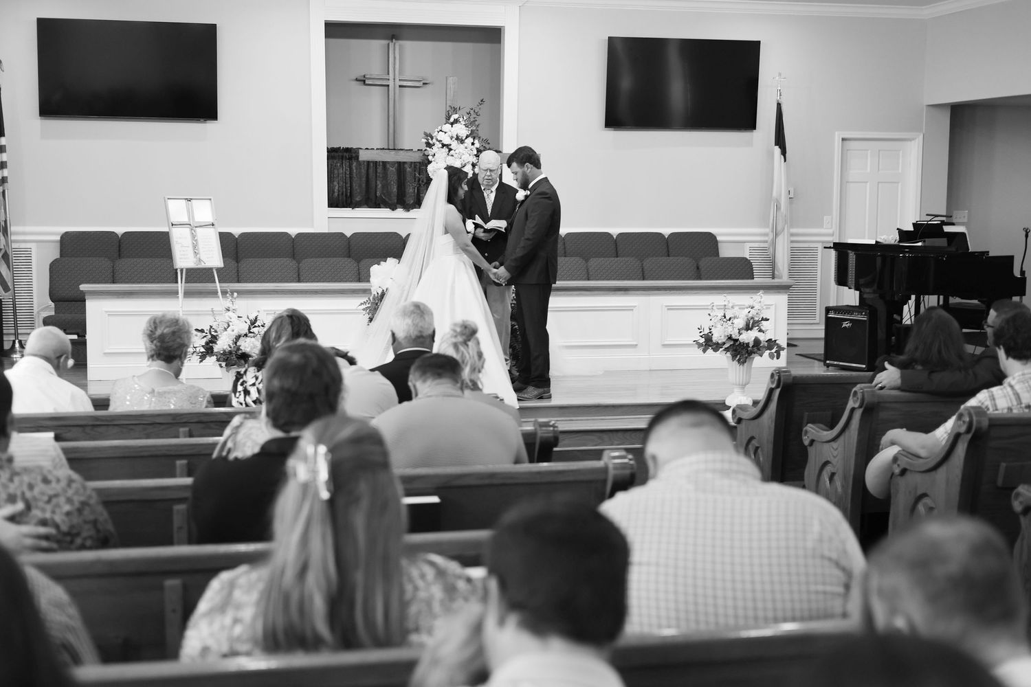 Black and white photo of a couple exchanging vows at a church wedding ceremony with guests seated in wooden pews.