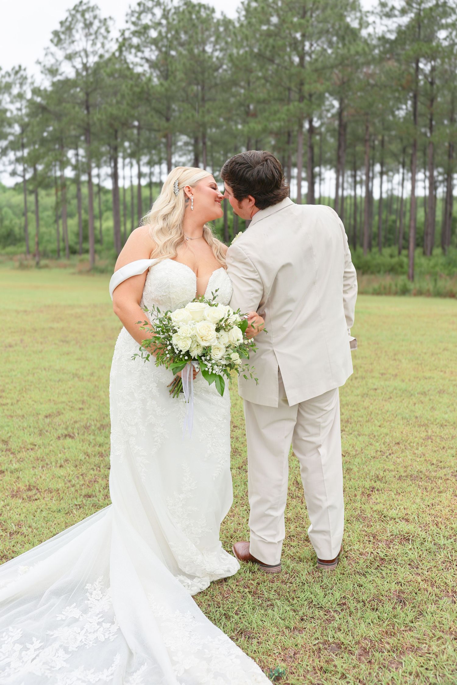 Bride and groom share a romantic nose-to-nose moment outdoors, bride holding white floral bouquet in lace gown with train.