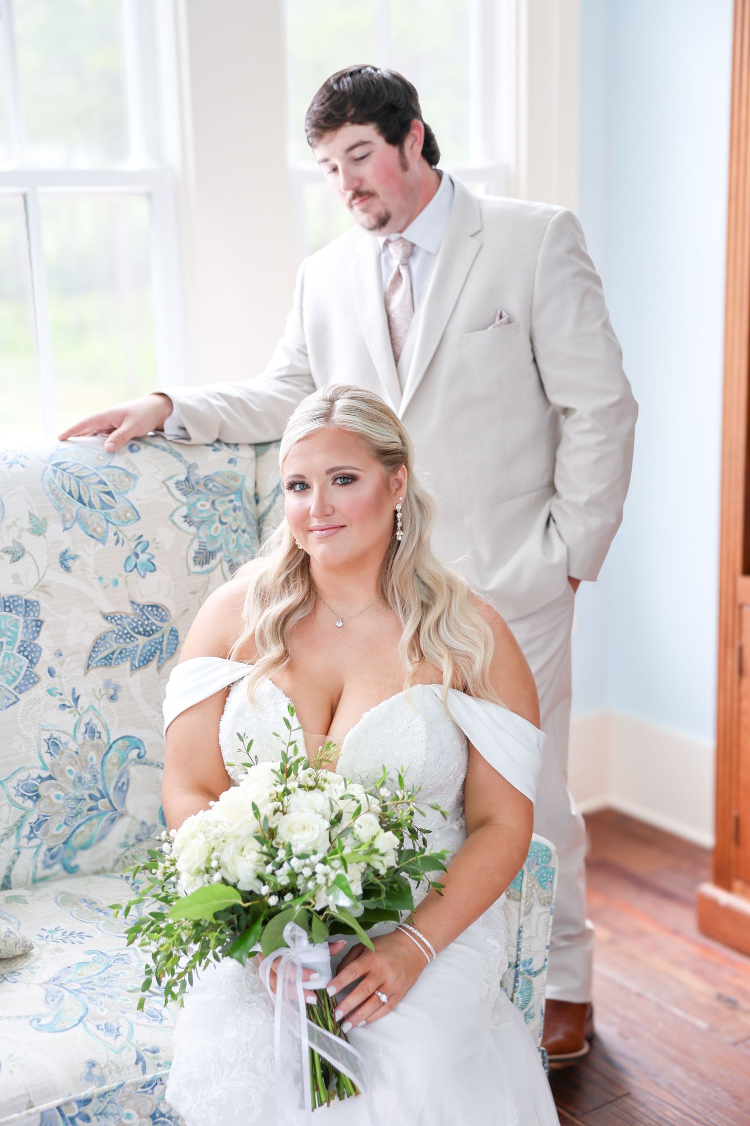 Bride in off-shoulder lace gown holds white bouquet seated as groom in tan suit stands behind her indoors.