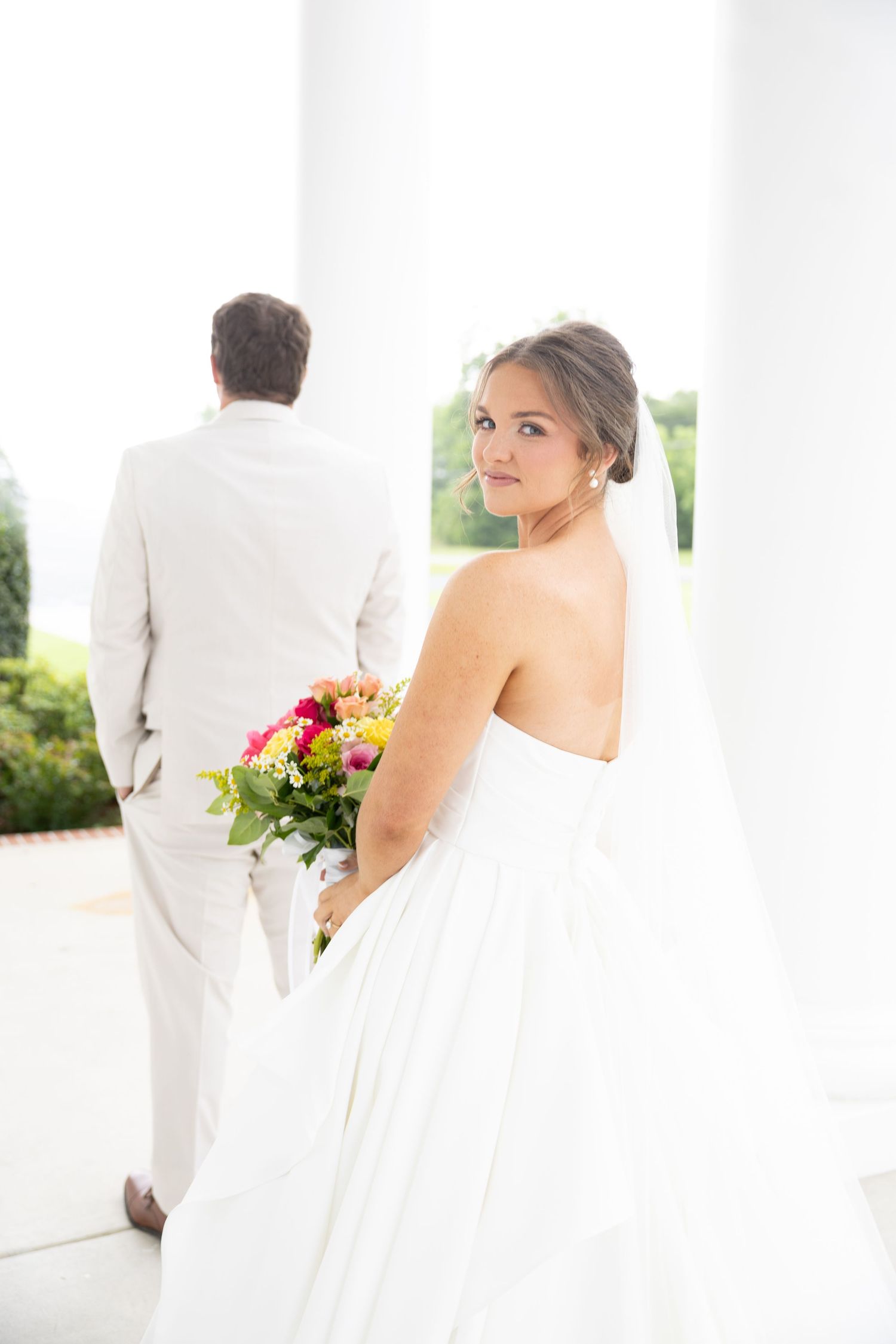 Bride in white strapless gown holds colorful bouquet, glancing back as groom in tan suit stands near white columns.