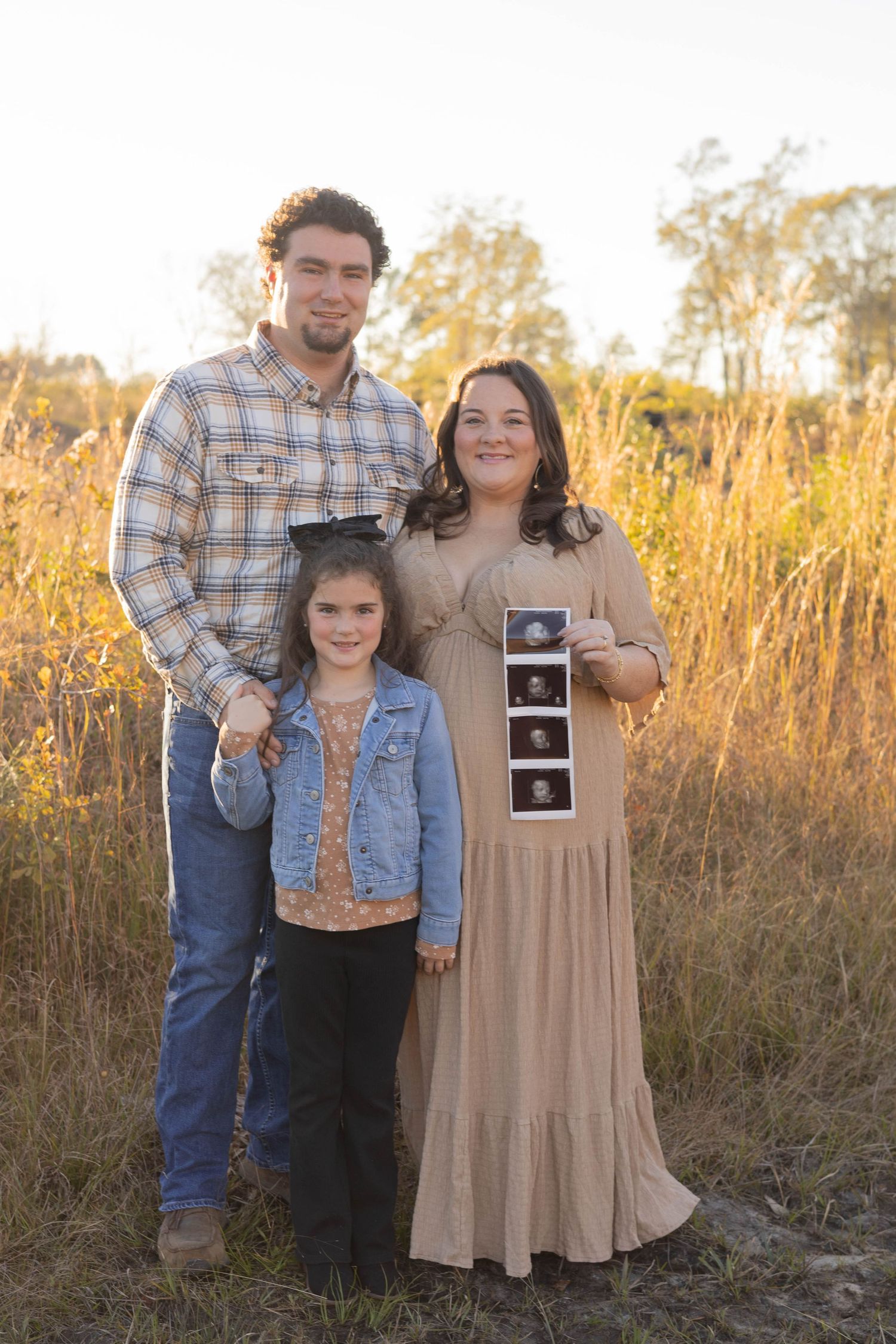 A couple and young girl pose outdoors holding ultrasound photos, announcing a pregnancy in a golden field at sunset.