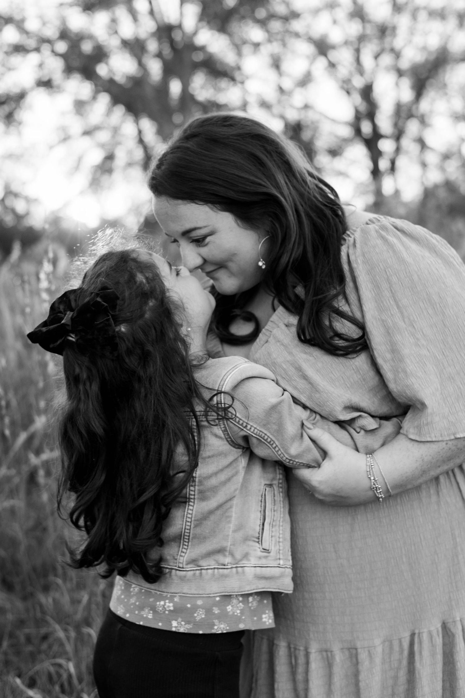 Black and white photo of a mother and young daughter sharing a tender kiss outdoors among tall grasses.
