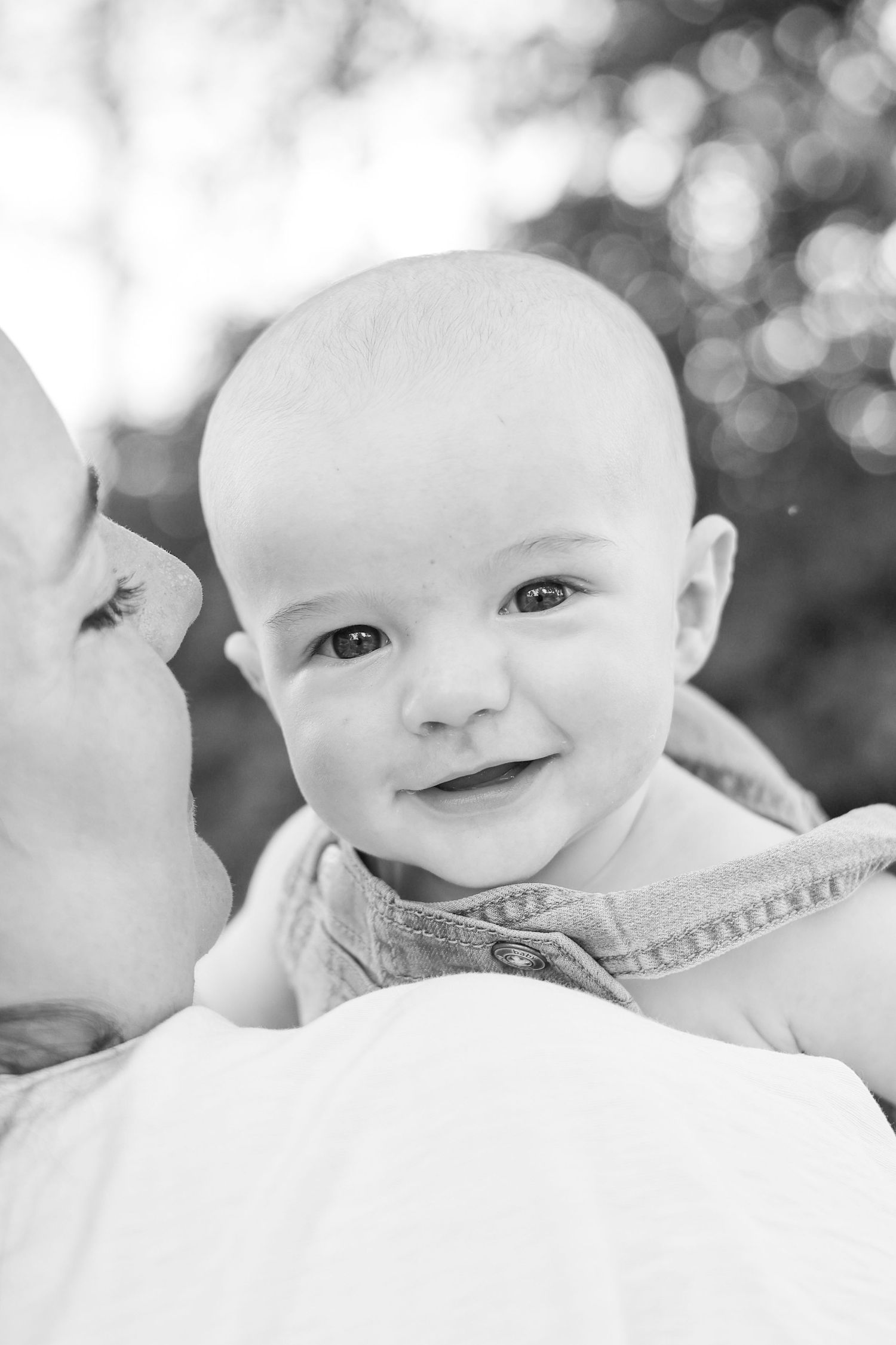 Black and white photo of a smiling baby being kissed by mother outdoors with bokeh background.