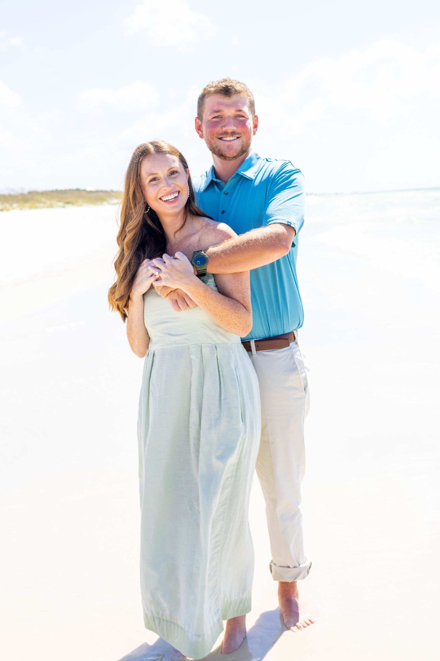 Happy couple embracing on a sunny beach, woman in mint dress, man in blue polo shirt, smiling outdoors.