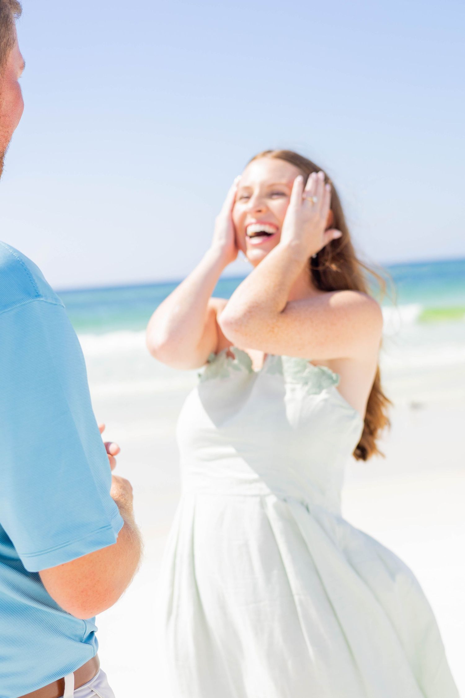 Joyful woman in white dress laughing on sunny beach with man in blue shirt during romantic proposal.