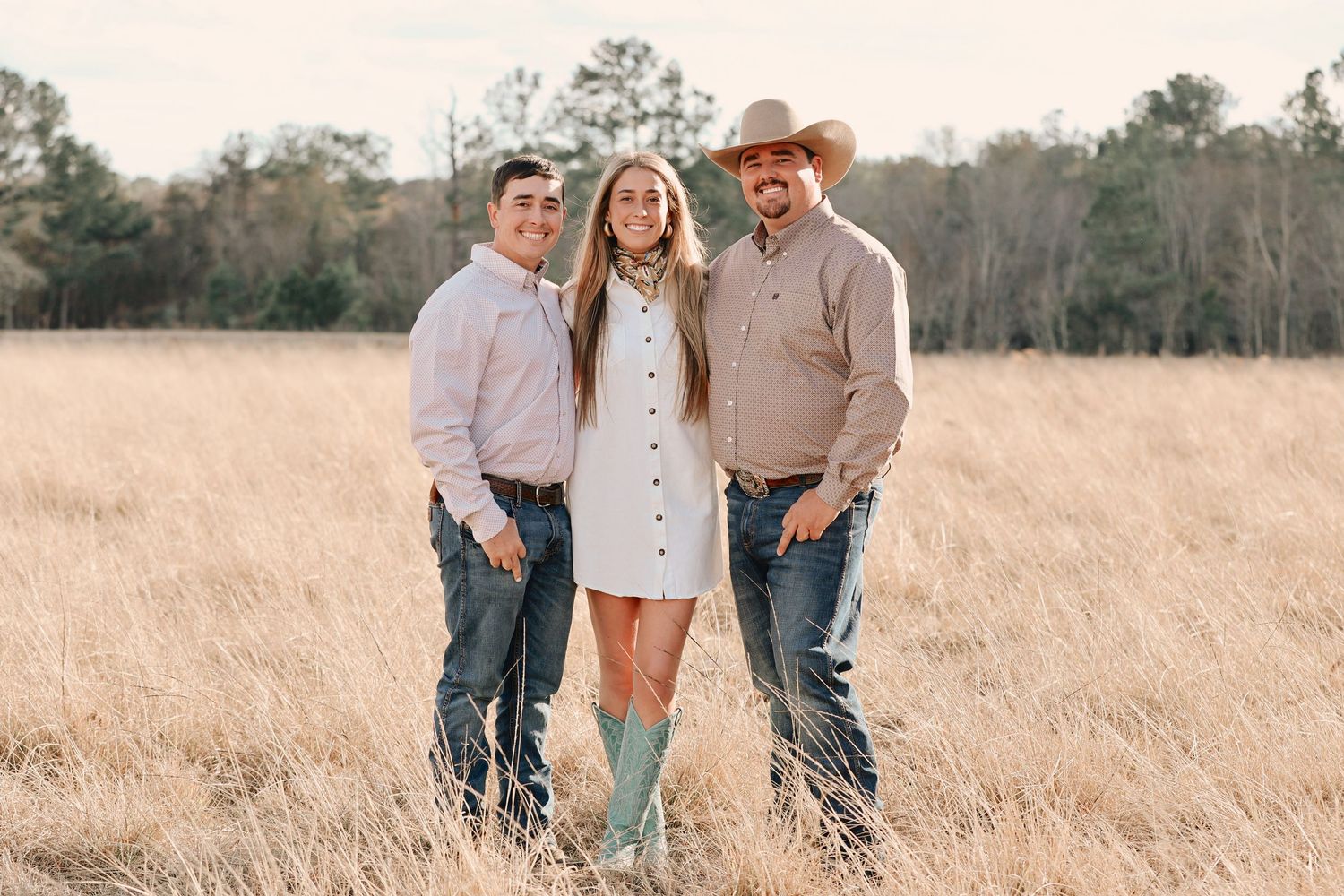 Three people smiling in a golden field, woman in white dress with green boots flanked by two men in casual Western attire.