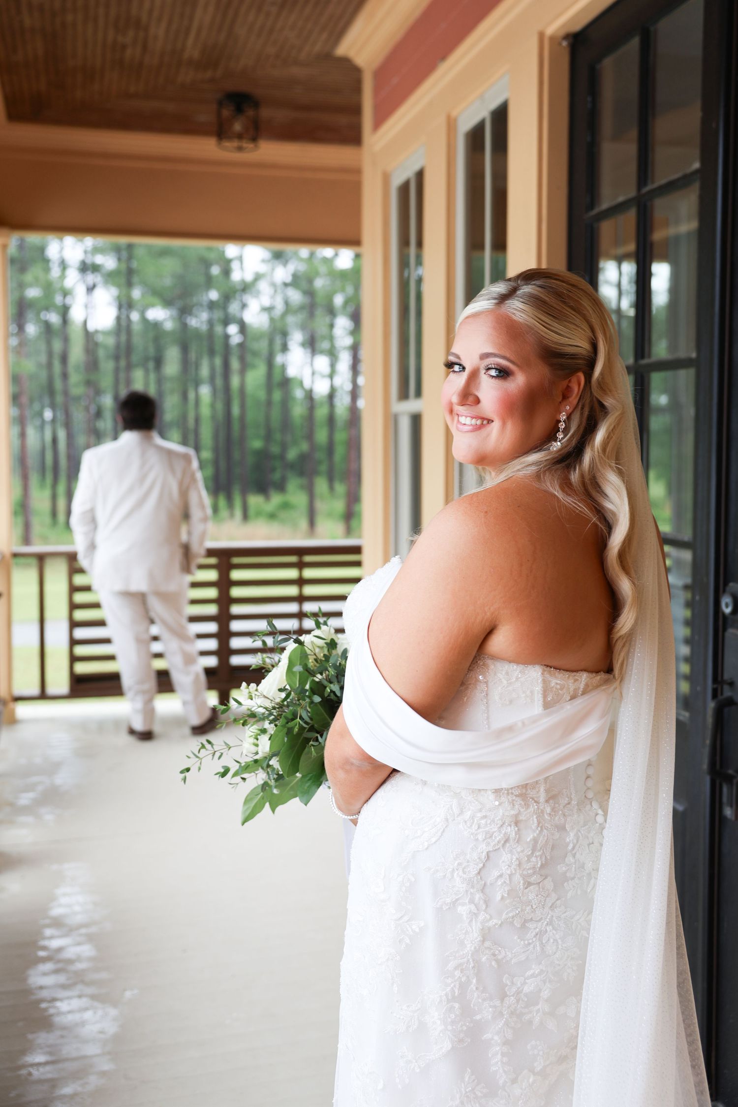 Bride in strapless lace gown holding greenery bouquet smiles on porch while groom stands with back turned.