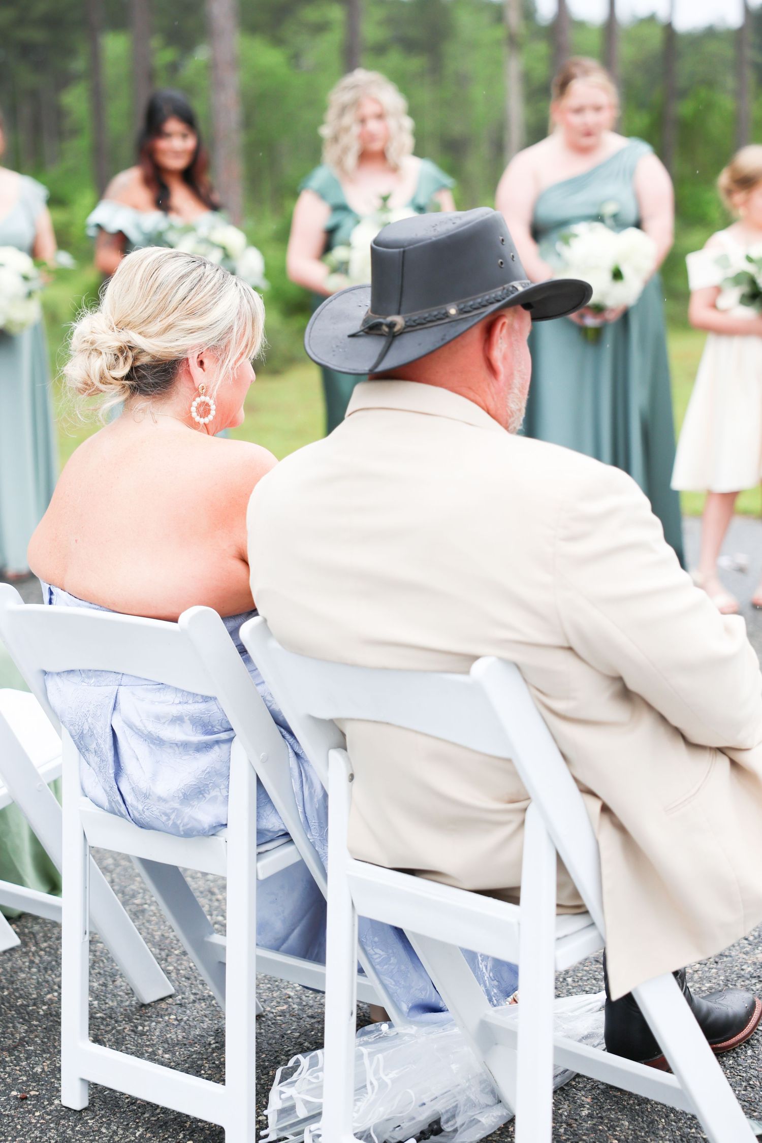 Father of bride in tan suit and cowboy hat seated with wife watching bridesmaids in sage green.
