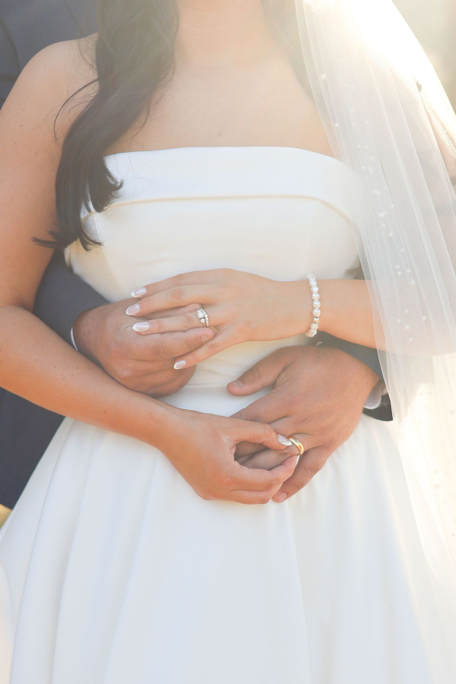 Close-up view of hands wearing a diamond engagement ring against a white wedding dress background.