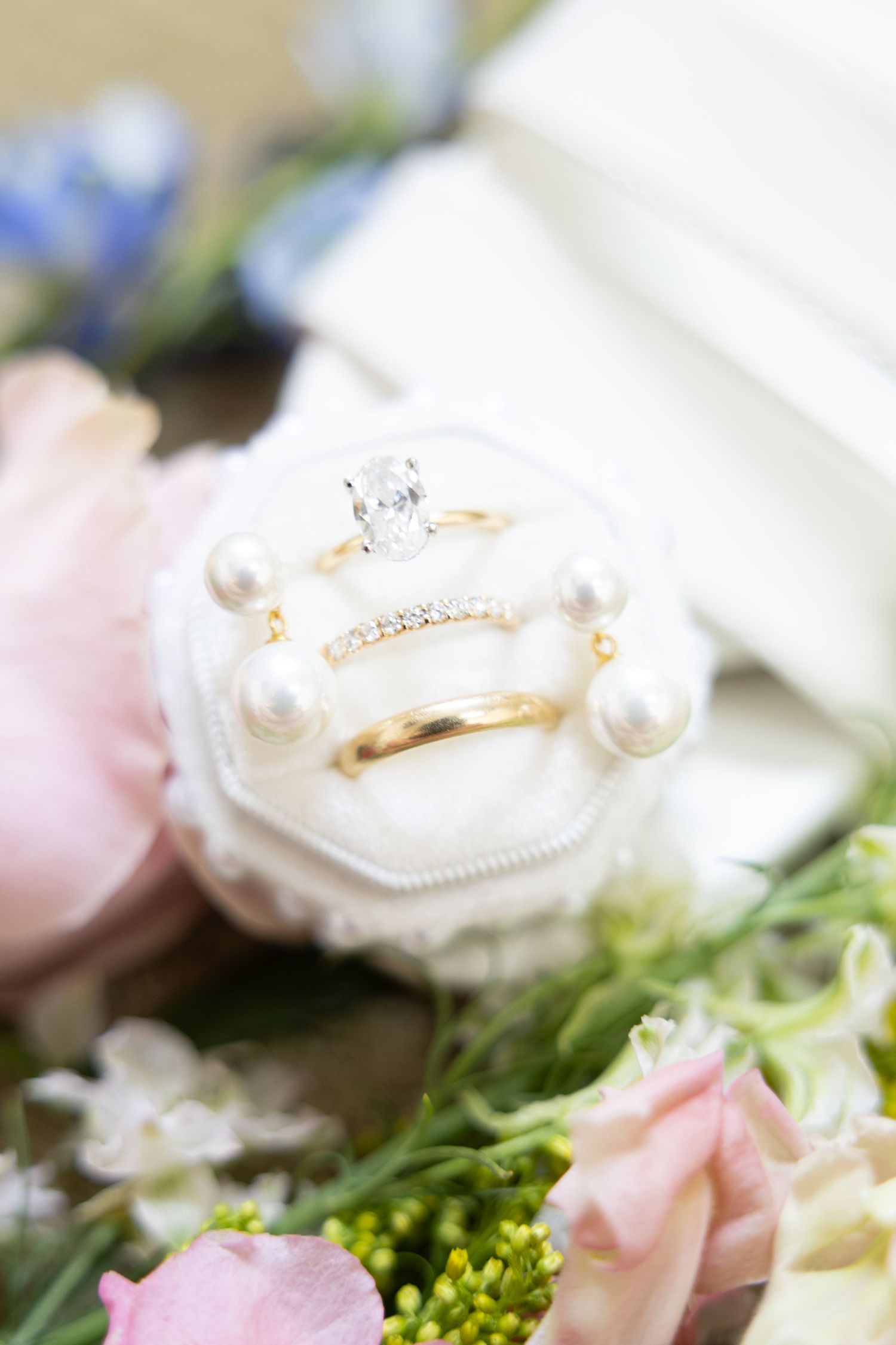 Gold wedding rings with diamonds displayed in a pearl-adorned velvet ring box surrounded by pink and white flowers.