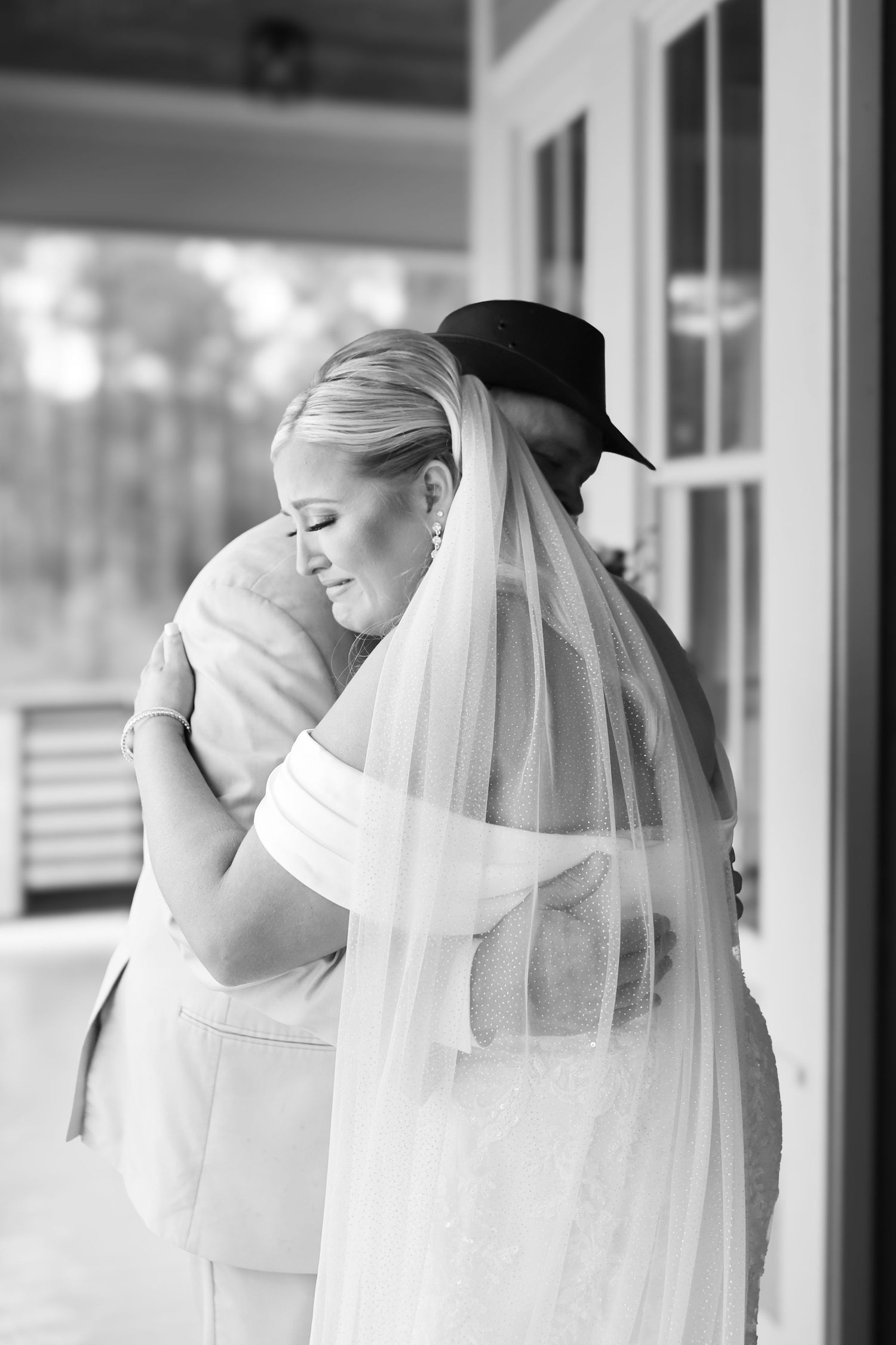 Black and white photo of a bride in a veil embracing a man in a cowboy hat on a porch.