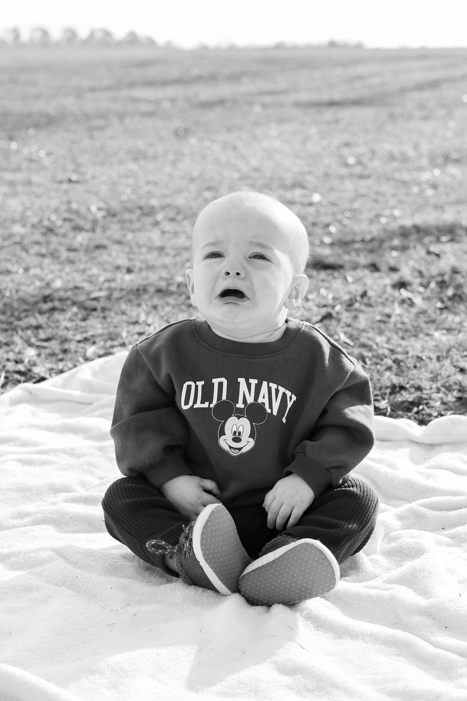 Black and white photo of a crying baby in an Old Navy Mickey Mouse sweatshirt sitting on a blanket outdoors.
