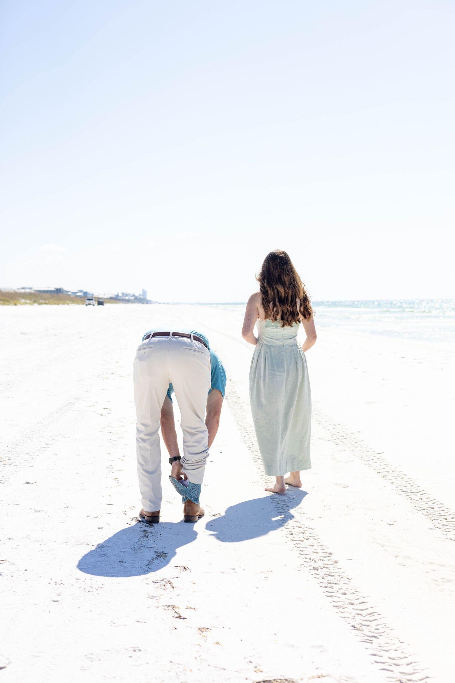 Couple walking on white sand beach, man bending down while woman in mint dress stands nearby with ocean in background.