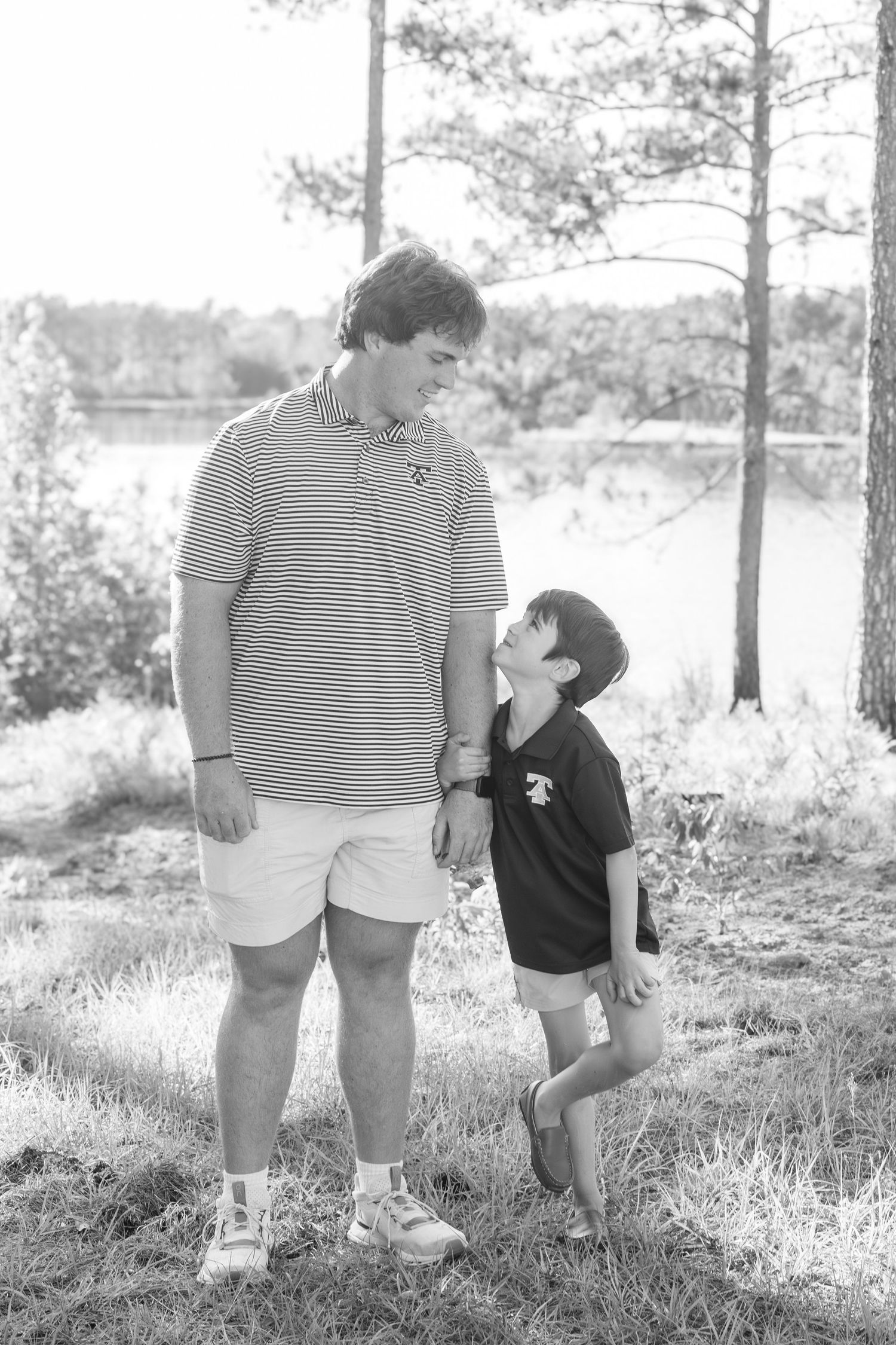 Black and white photo of a tall teen and young boy smiling at each other outdoors near a lake.