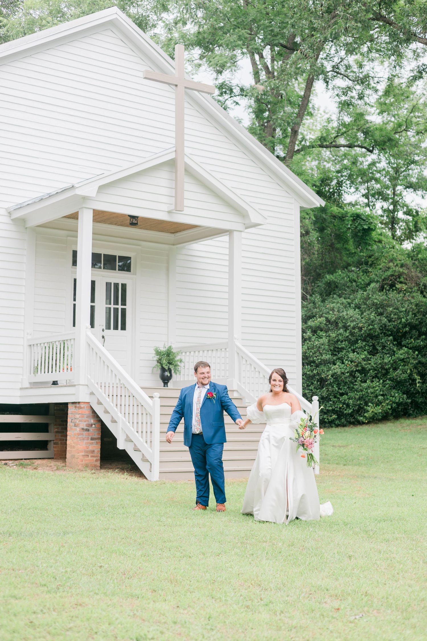 Bride and groom holding hands walking from white chapel with cross at outdoor wedding venue.