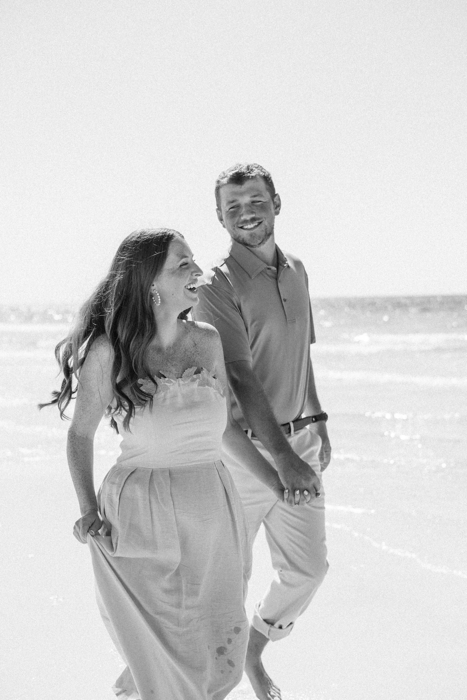Black and white photo of laughing couple walking hand in hand along the ocean shoreline.