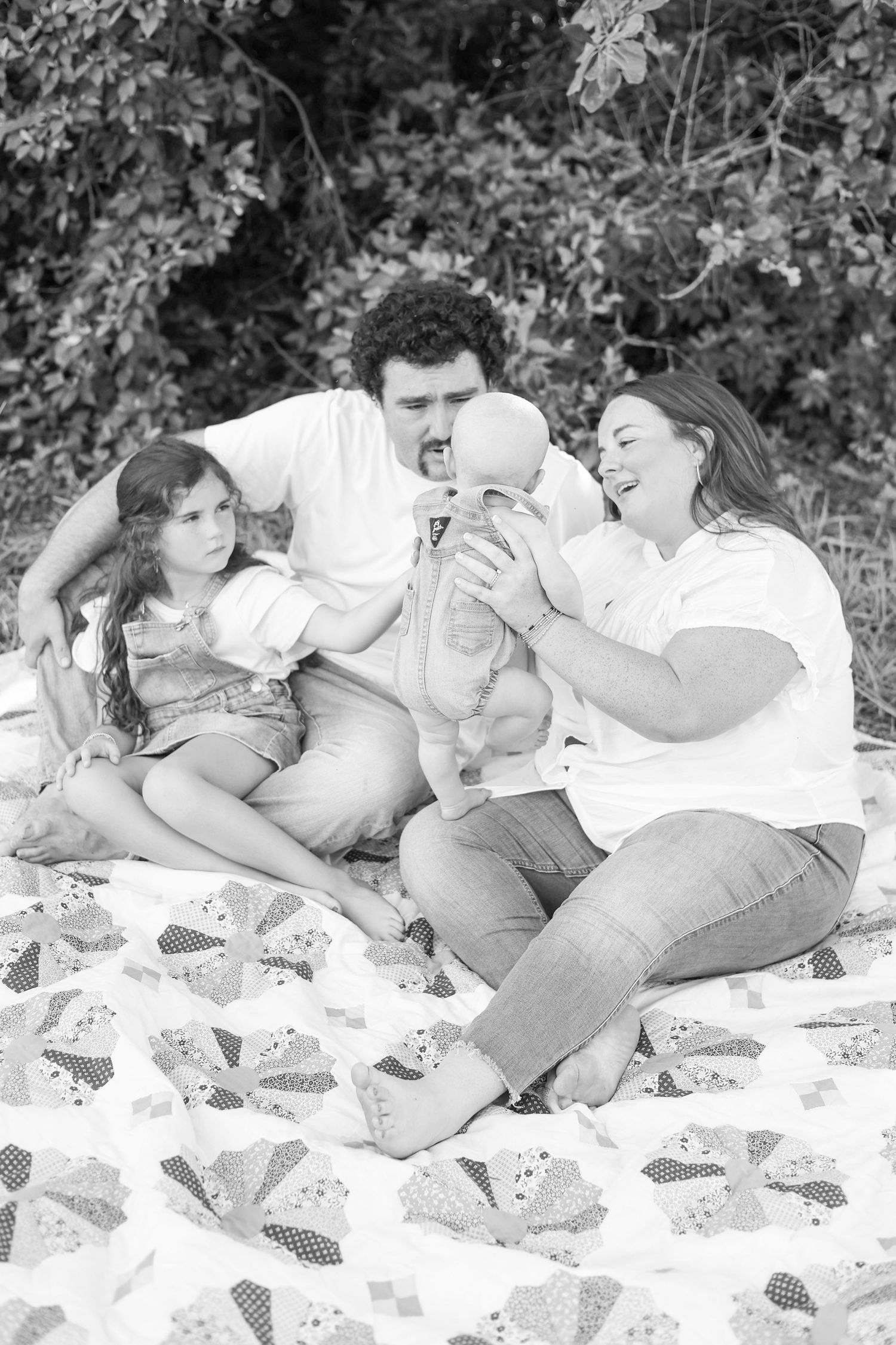 Black and white family photo of parents with young daughter and baby sitting on quilt outdoors near lush greenery.